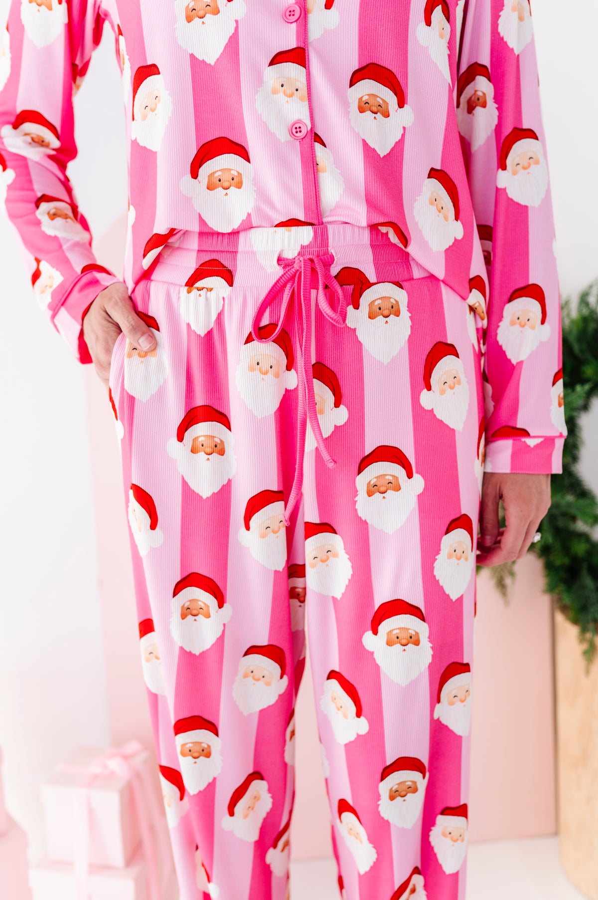 A close-up photo of a woman wearing pink Santa Claus bamboo pajamas, standing in a bright white room with a green wreath and wrapped gifts in the background.