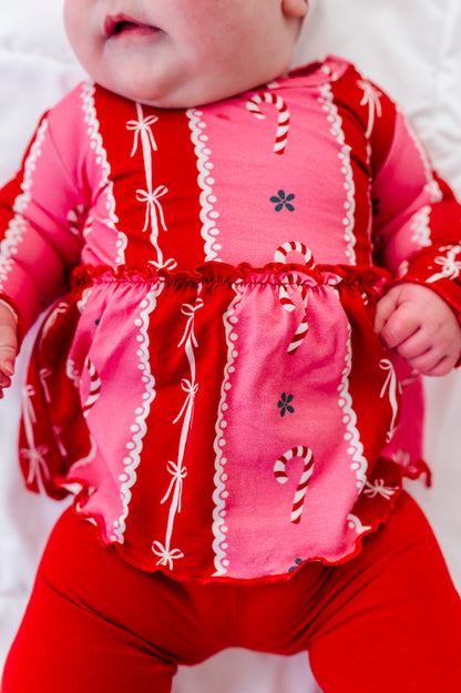A close-up of a baby wearing a festive red and pink long-sleeve bamboo top with white lace trim, candy cane prints, and red leggings, lying on a white background.