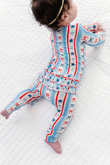 Baby in a patriotic bamboo zip romper with red, white, and blue stripes, bows, and stars on a white textured surface