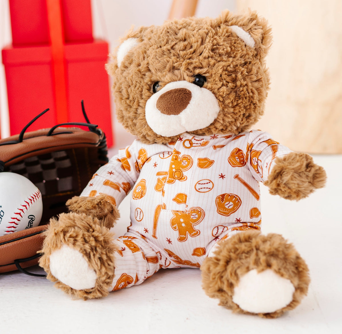 A brown plush teddy bear wearing a white and orange baseball-themed onesie sits next to a baseball glove with a baseball, with red gift boxes and a wooden bat in the background.