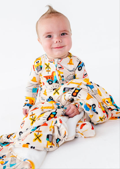 A baby sitting on a white studio background wearing a long-sleeve footed sleeper with a colorful construction vehicle print.