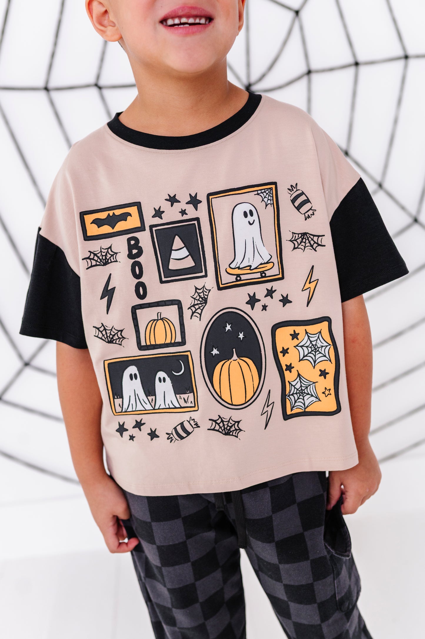 A child wearing a short-sleeve t-shirt with a beige body and black sleeves, decorated with a Halloween patchwork graphic of pumpkins, ghosts, bats, and spiderwebs, paired with black and gray checkered pants.