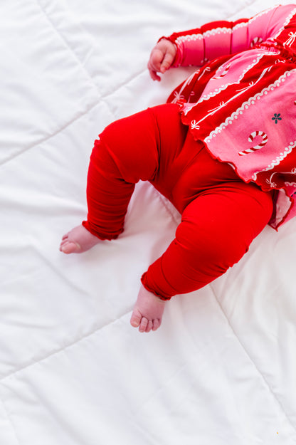 A baby lying on a white quilted blanket, wearing bright red leggings and a pink and red holiday-themed long-sleeve bamboo top with candy cane and snowflake details.
