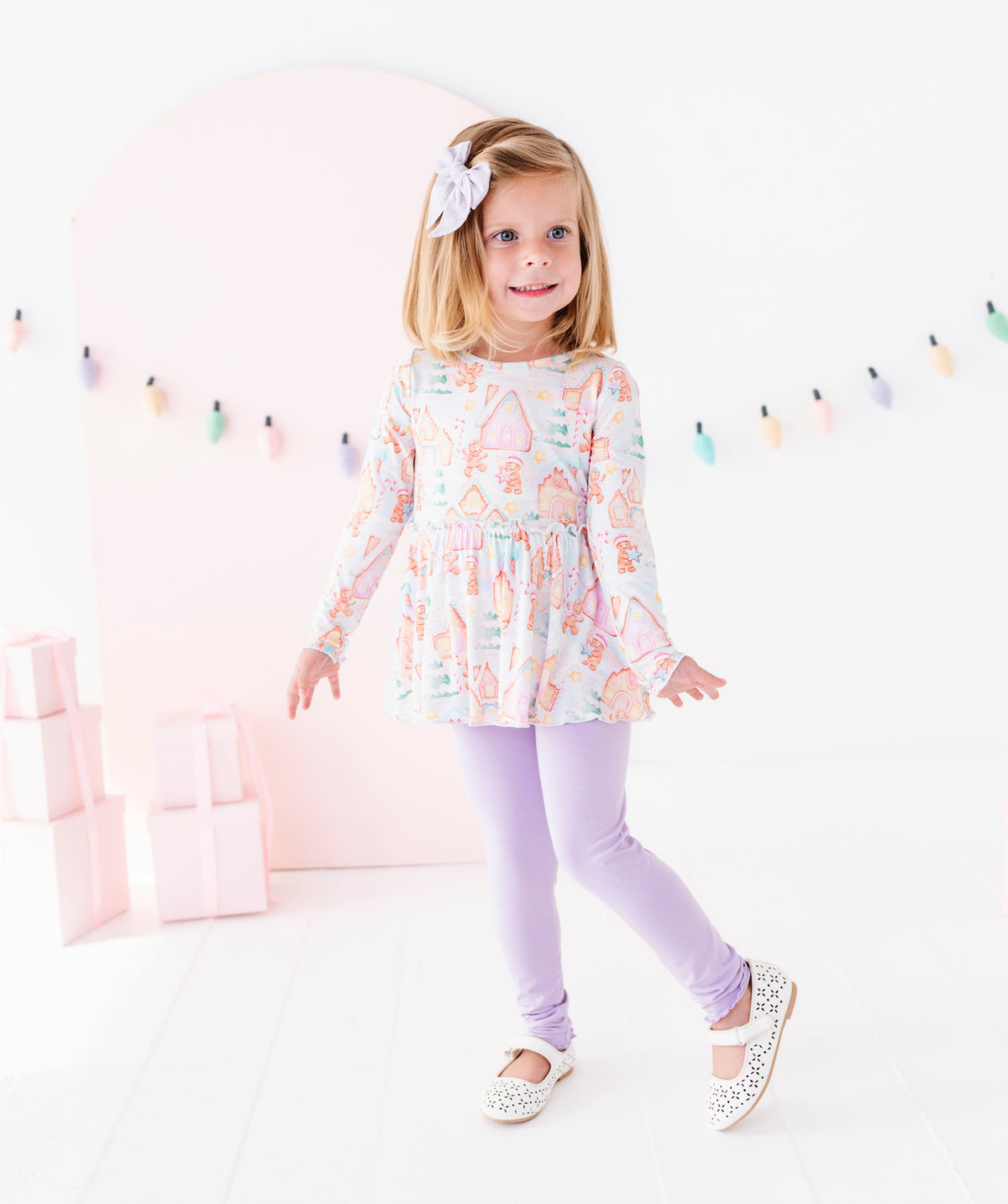 A young girl with blonde hair wearing a pastel multicolor patterned long-sleeve tunic top, lavender leggings, and white Mary Jane shoes, standing in a bright white studio with a pink arch background.