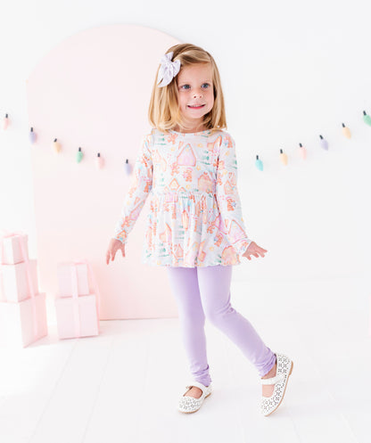A young girl with blonde hair wearing a pastel multicolor patterned long-sleeve tunic top, lavender leggings, and white Mary Jane shoes, standing in a bright white studio with a pink arch background.