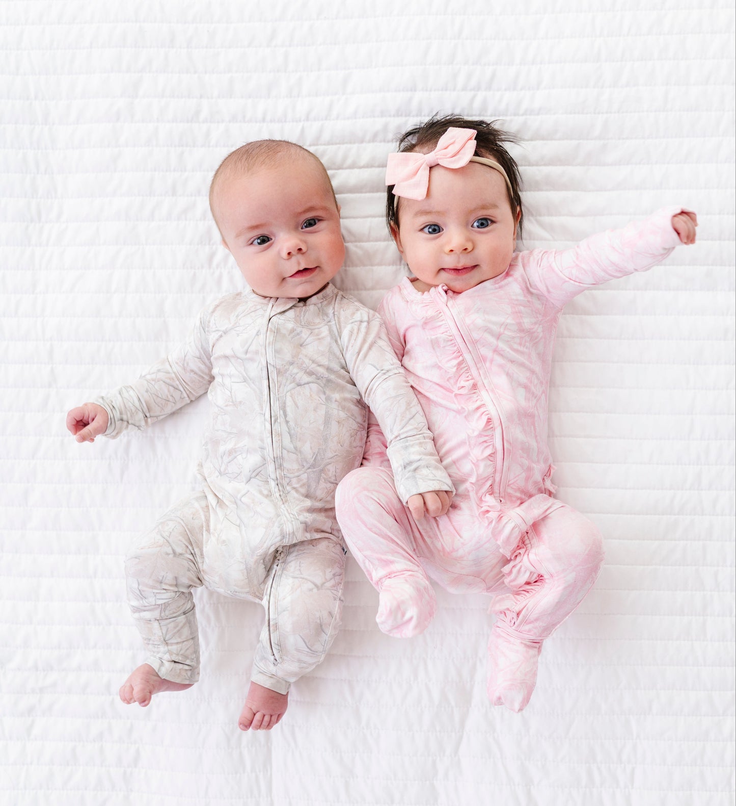 Two babies in coordinating tree camo rompers  lying on a white textured surface