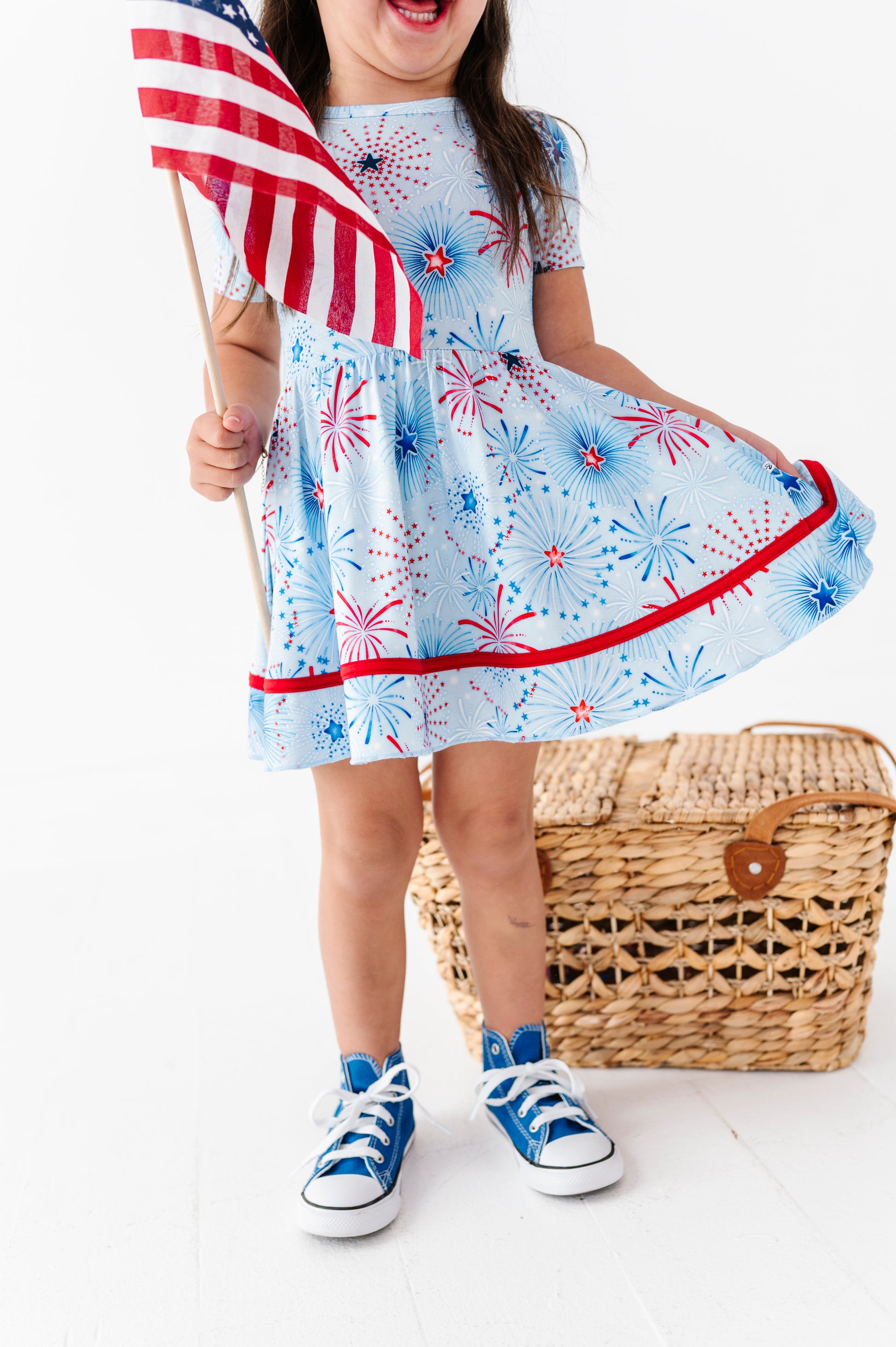 Child wearing a bamboo dress with fireworks pattern, holding an American flag, standing next to a woven basket on a white background.