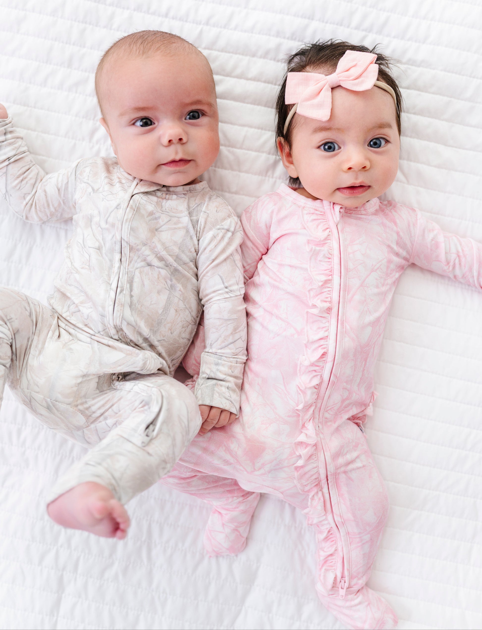 Two babies lying on a white blanket, one in a beige outfit and the other in a pink outfit with a bow.