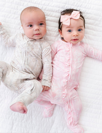 Two babies lying on a white blanket, one in a beige outfit and the other in a pink outfit with a bow.