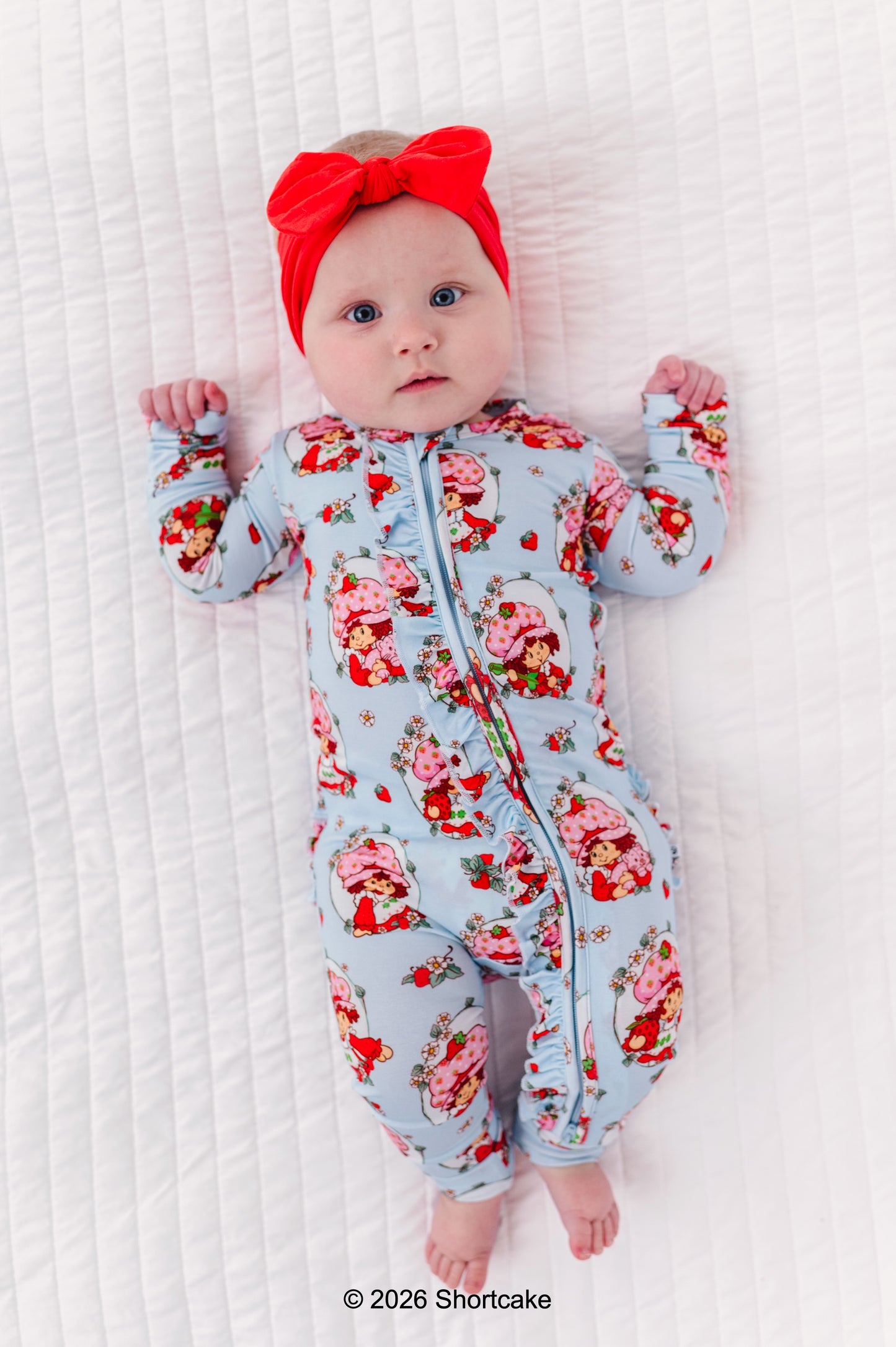 Baby wearing a Strawberry Shortcake™ bamboo onesie and red headband on a white textured surface