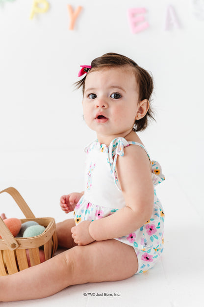 Baby in a floral PEEPS® romper sitting next to a basket of Easter eggs on a white background