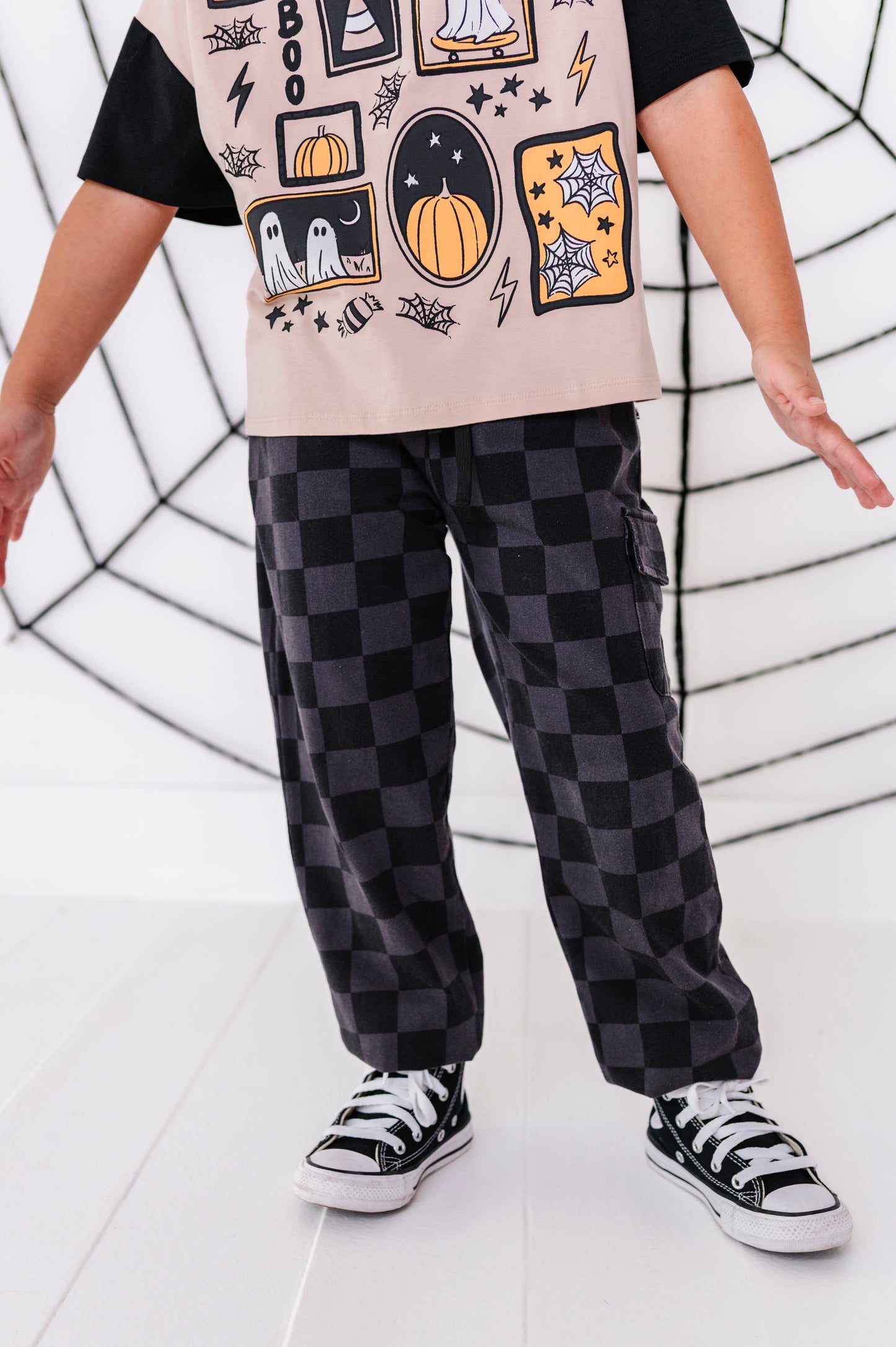 A child wearing black and dark gray checkered cargo pants, a black t-shirt with a beige graphic print, and black canvas sneakers, standing in front of a white wall with a black spiderweb design.