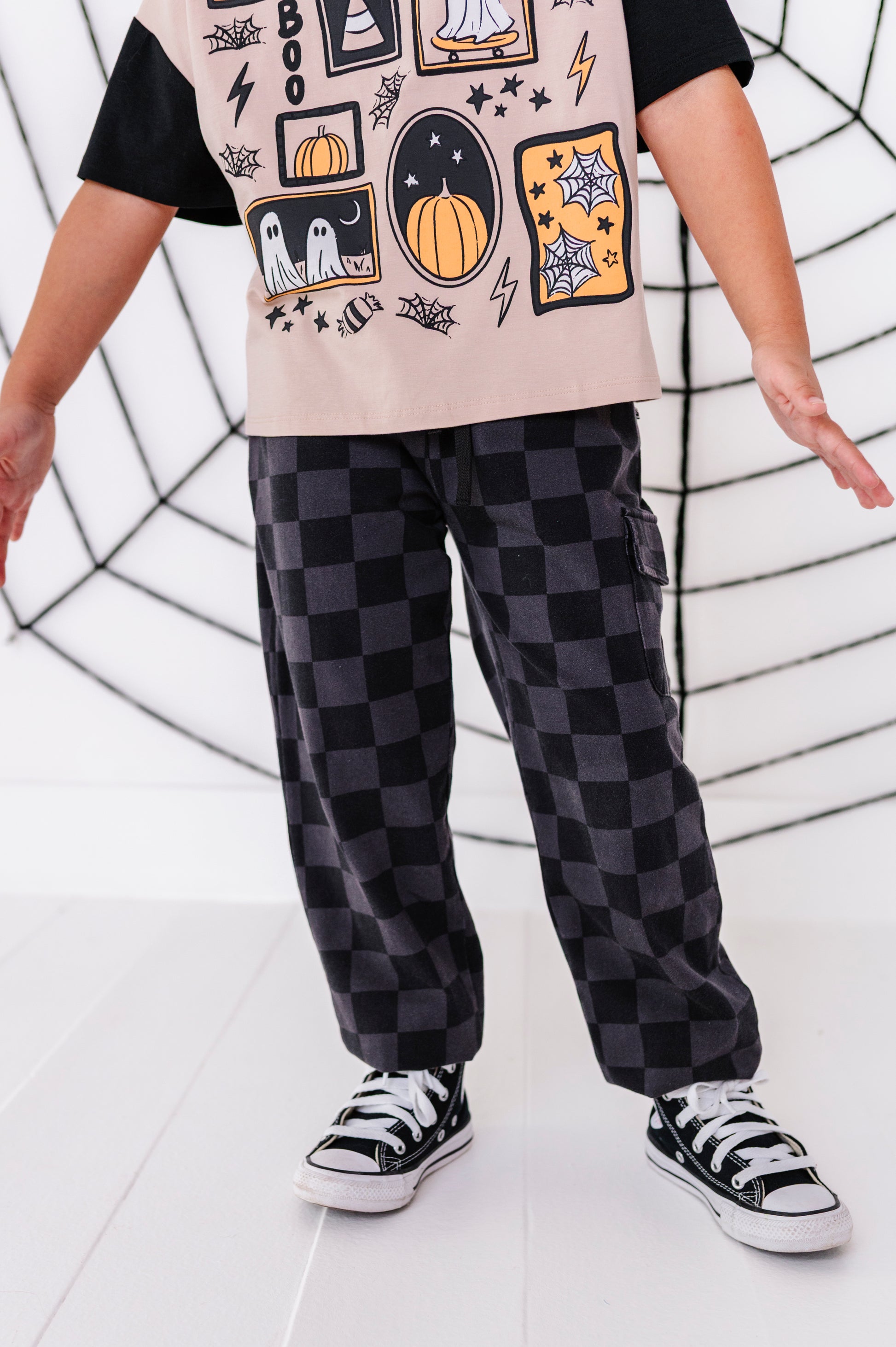 A child wearing black and dark gray checkered cargo pants, a black t-shirt with a beige graphic print, and black canvas sneakers, standing in front of a white wall with a black spiderweb design.