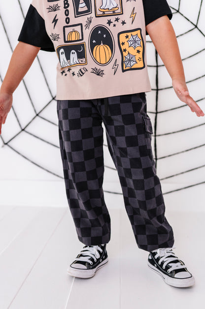 A child wearing black and dark gray checkered cargo pants, a black t-shirt with a beige graphic print, and black canvas sneakers, standing in front of a white wall with a black spiderweb design.
