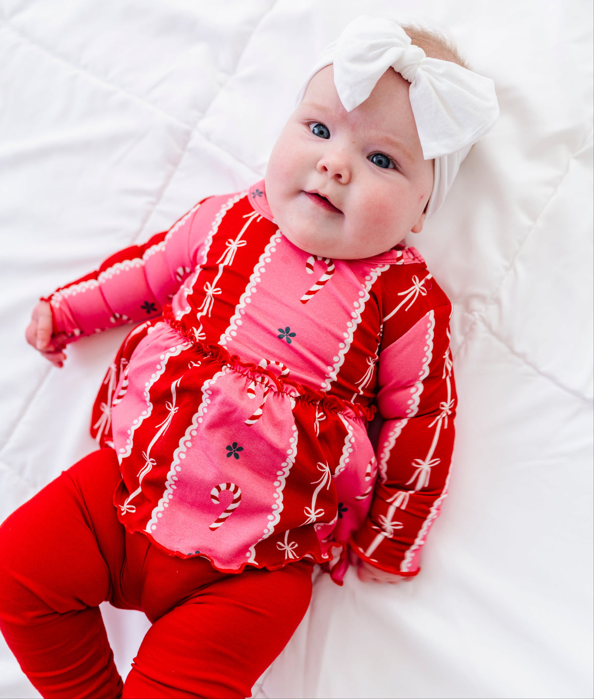 A baby lying on a white quilted blanket, wearing a festive red and pink holiday-themed long-sleeve bamboo top with white trim, matching red leggings, and a white bow headband.