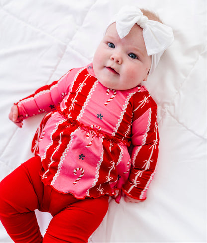 A baby lying on a white quilted blanket, wearing a festive red and pink holiday-themed long-sleeve bamboo top with white trim, matching red leggings, and a white bow headband.