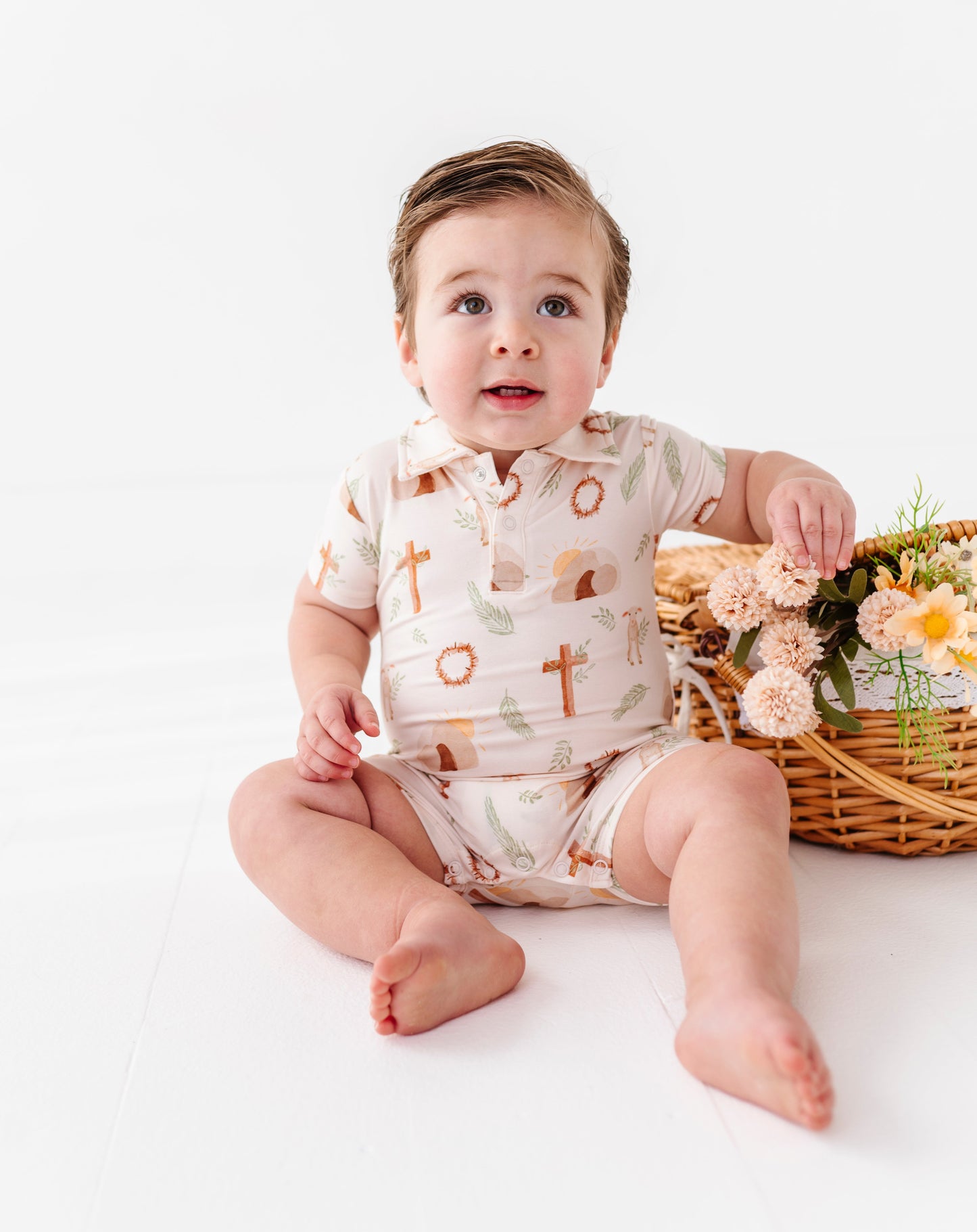Baby sitting on a white surface wearing a bamboo Easter onesie featuring the cross, crown of thorns, rolled-away stone, etc. with a basket of flowers in the background