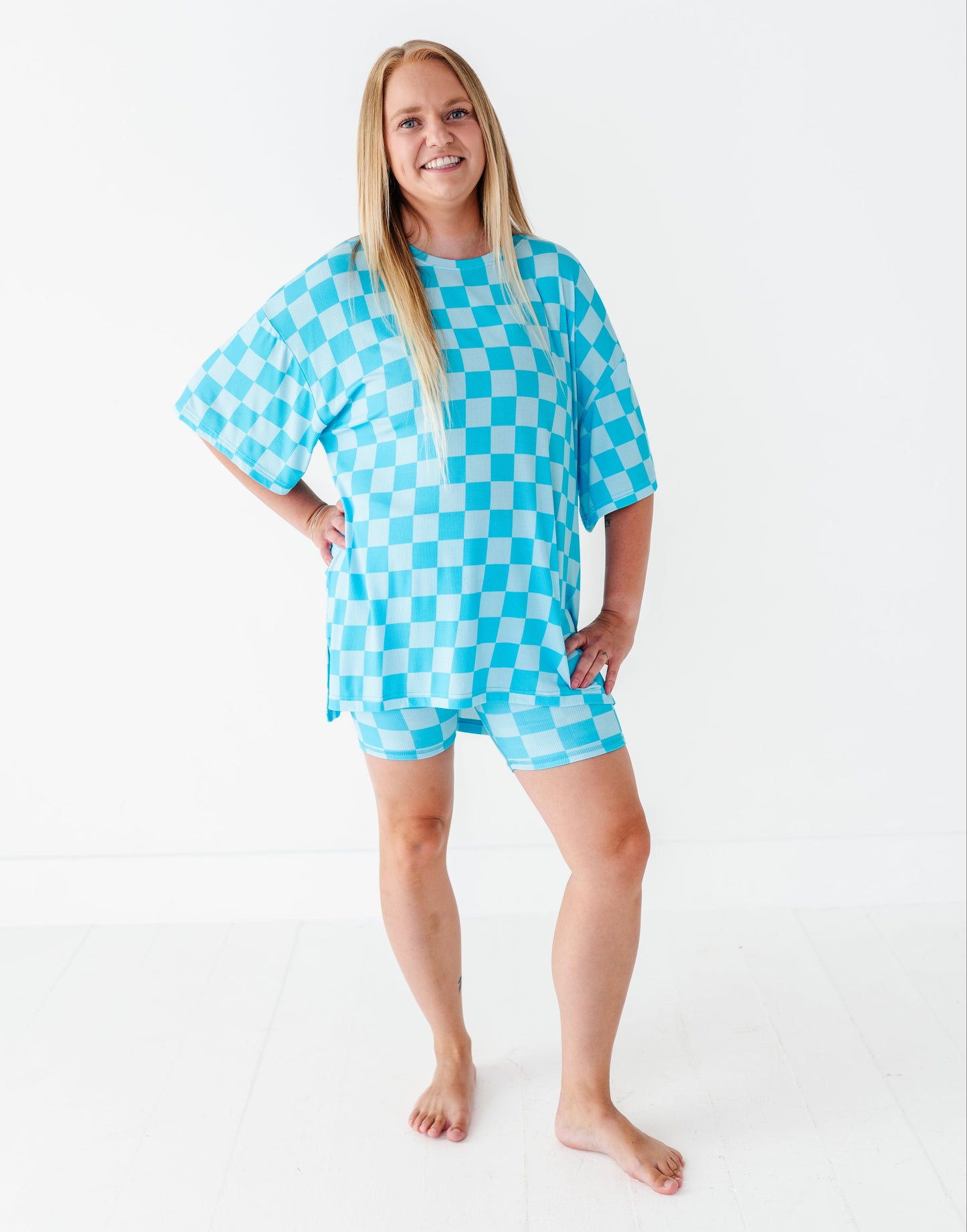 A woman modeling a matching two-piece lounge set featuring a bright turquoise and white checkerboard pattern, consisting of an oversized short-sleeve top and matching shorts, set against a plain white studio background.