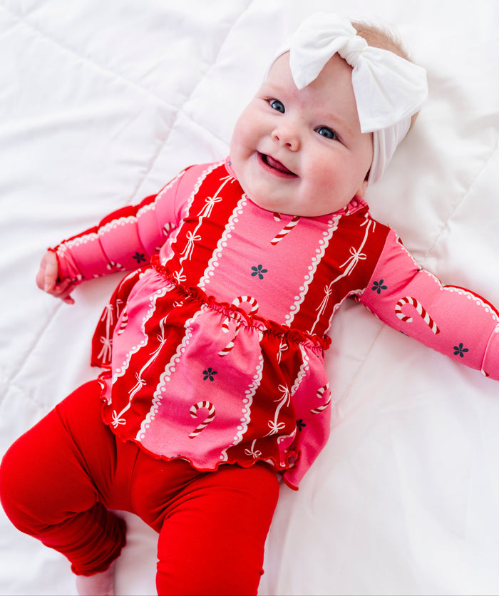 A smiling baby lying on a white quilted blanket, wearing a festive Christmas-themed bamboo peplum outfit with a pink long-sleeve top featuring red stripes, white lace trim, candy cane prints, and red leggings, along with a white bow headband.