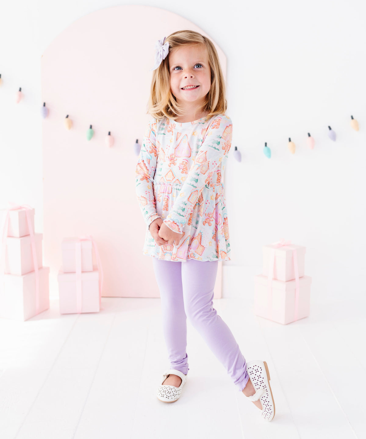 A little girl wearing a Christmas gingerbread-themed bamboo peplum top with purple leggings and white flats. She's standing in front of pink presents and a pastel Christmas lights banner. 
