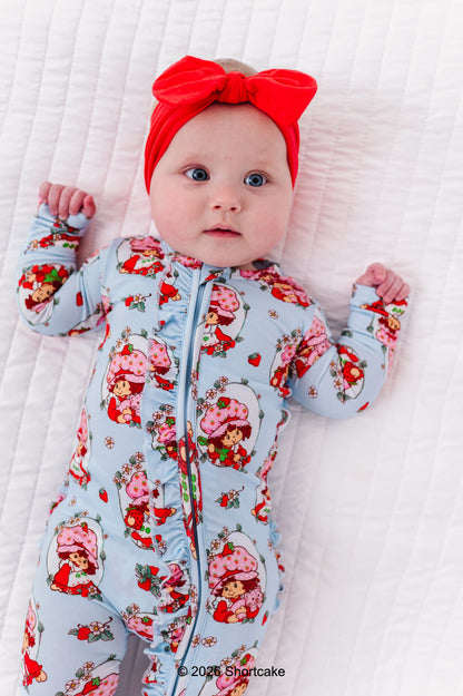 Baby wearing a Strawberry Shortcake™ bamboo zip romper and red headband on a white blanket