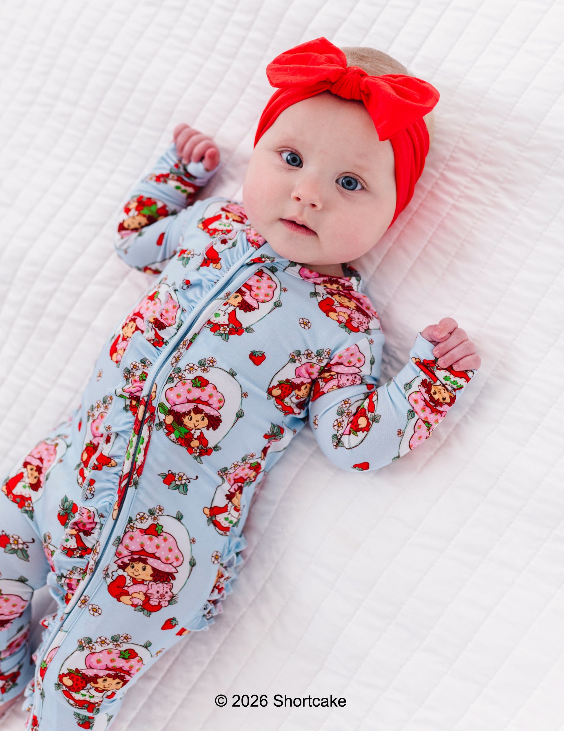 Baby wearing a bamboo Strawberry Shortcake™ zip romper and red headband on a white background