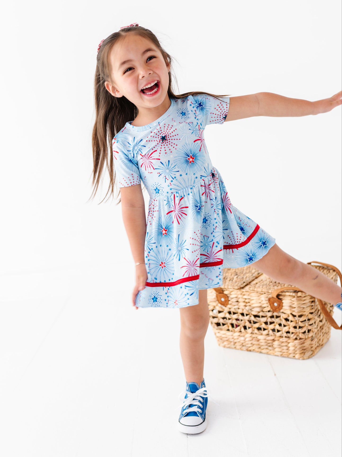 Young girl wearing a bamboo dress with red, white, and blue patriotic patterns, standing next to a woven bag on a white background.
