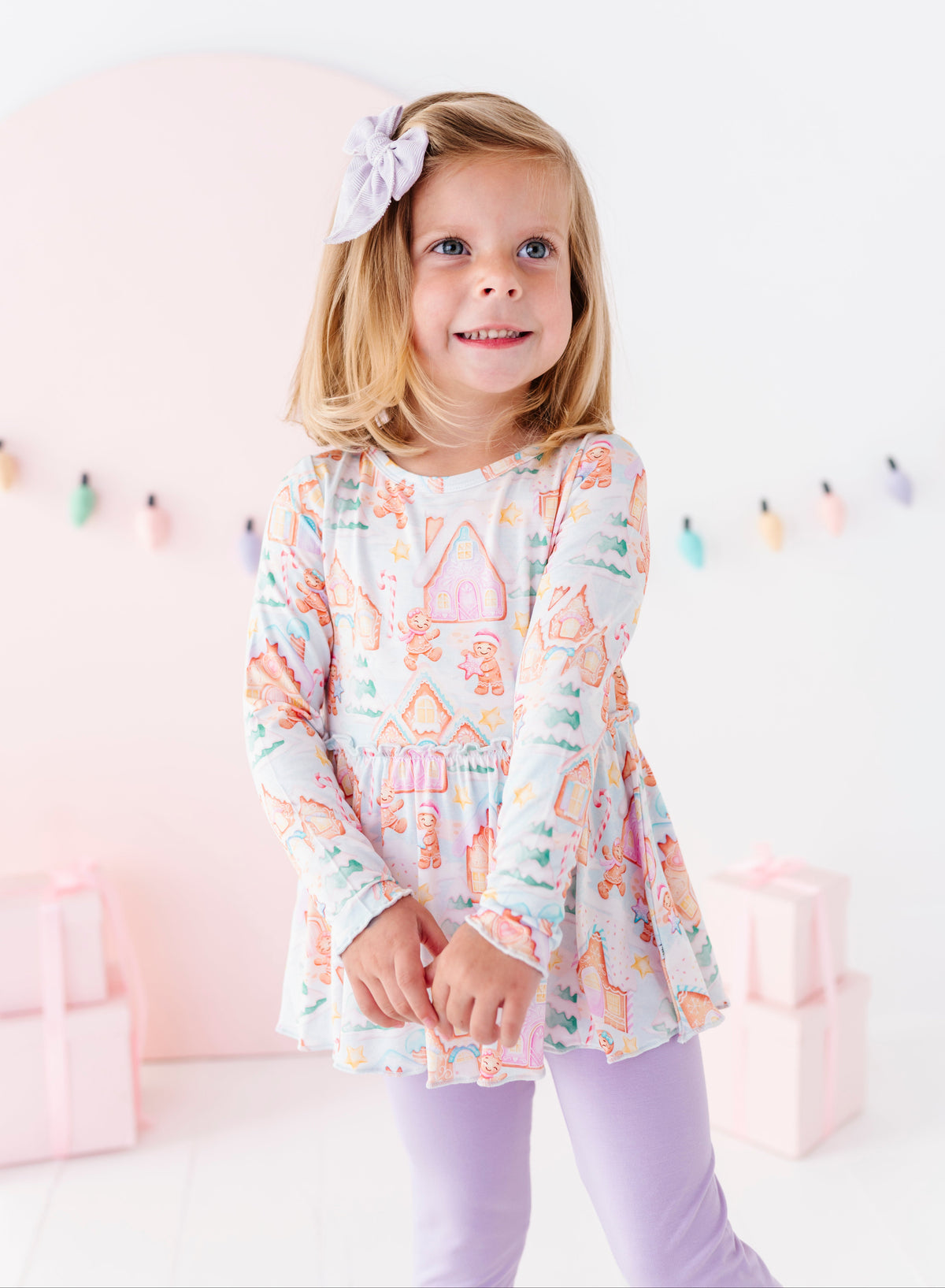 A little girl wearing a Christmas gingerbread-themed bamboo peplum top with purple leggings. She's standing in front of pink presents and a pastel Christmas lights banner. 