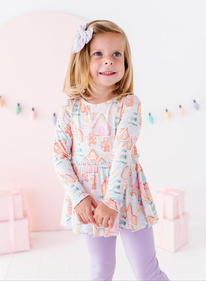 A little girl wearing a Christmas gingerbread-themed bamboo peplum top with purple leggings. She's standing in front of pink presents and a pastel Christmas lights banner. 