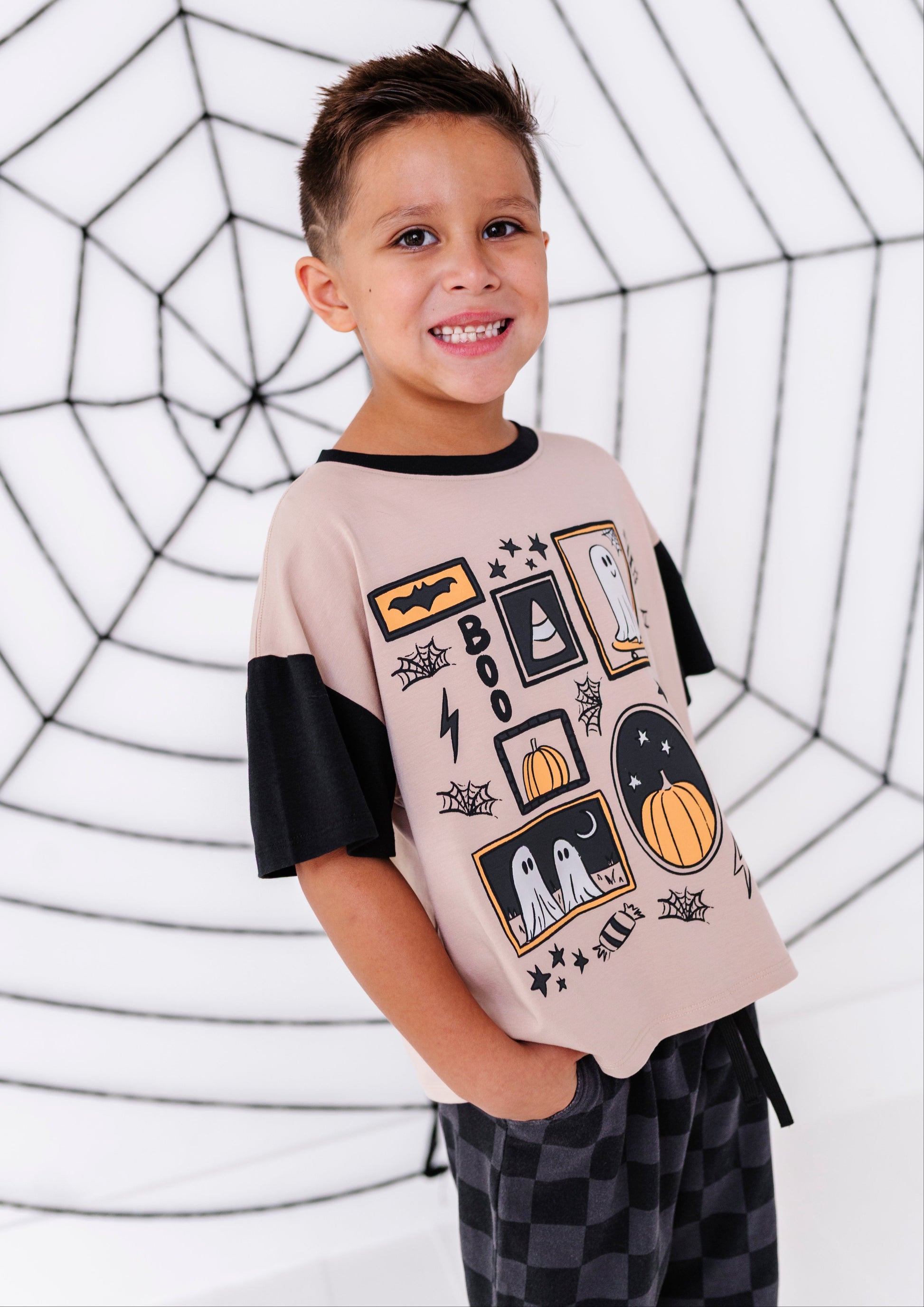A smiling young boy wearing a short-sleeve ringer t-shirt with a beige body, black sleeves, and a black and orange Halloween graphic print, paired with black and gray checkered pajama pants. He is standing in front of a white background decorated with a black spiderweb pattern.