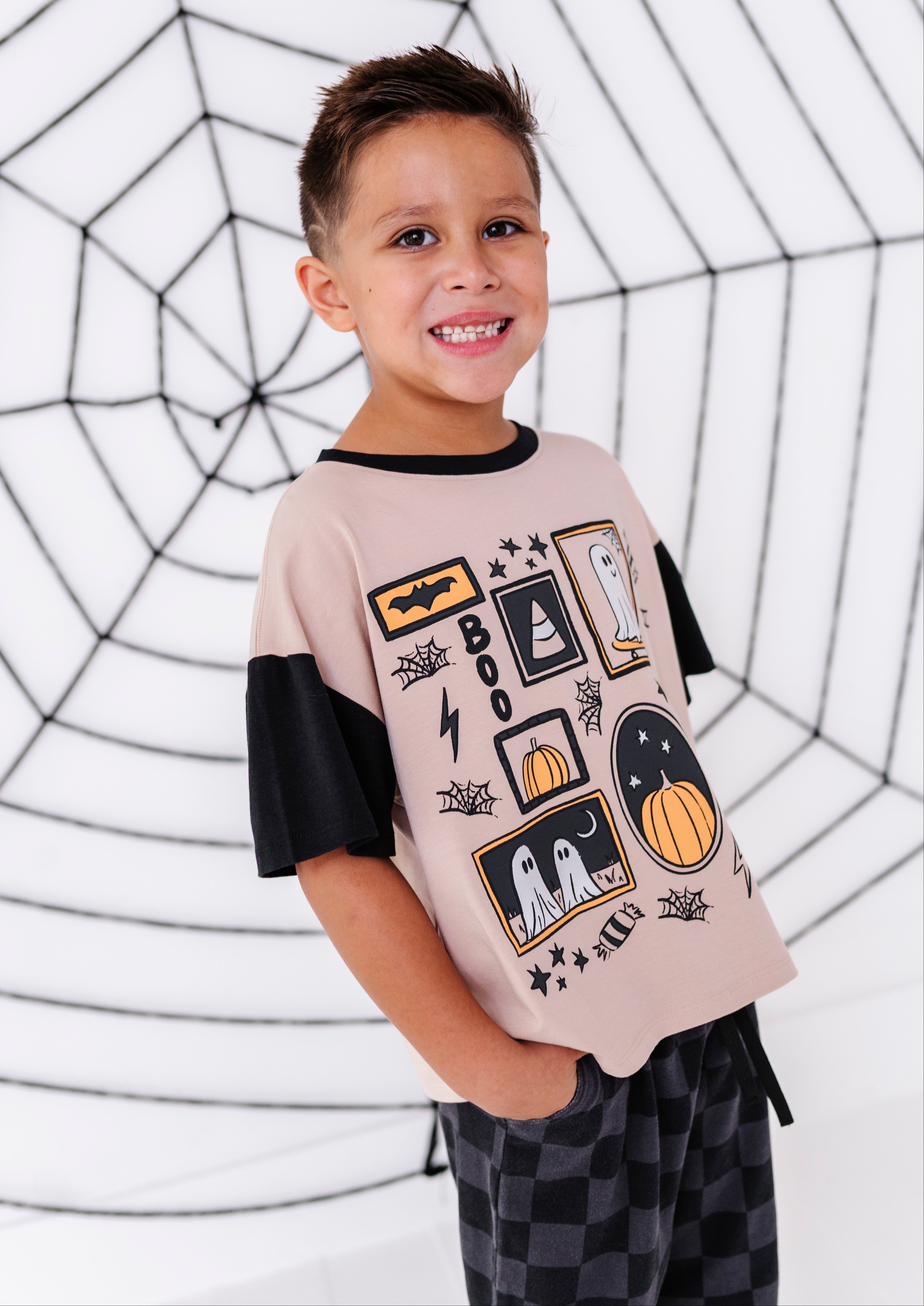 A smiling young boy wearing a short-sleeve ringer t-shirt with a beige body, black sleeves, and a black and orange Halloween graphic print, paired with black and gray checkered pajama pants. He is standing in front of a white background decorated with a black spiderweb pattern.