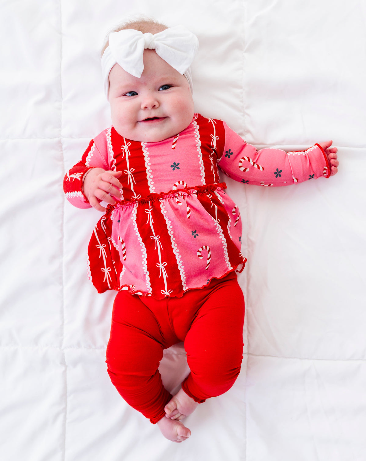A baby lying on a white quilted blanket, wearing a festive red and pink holiday-themed long-sleeve bamboo top with matching red pants and a white bow headband.
