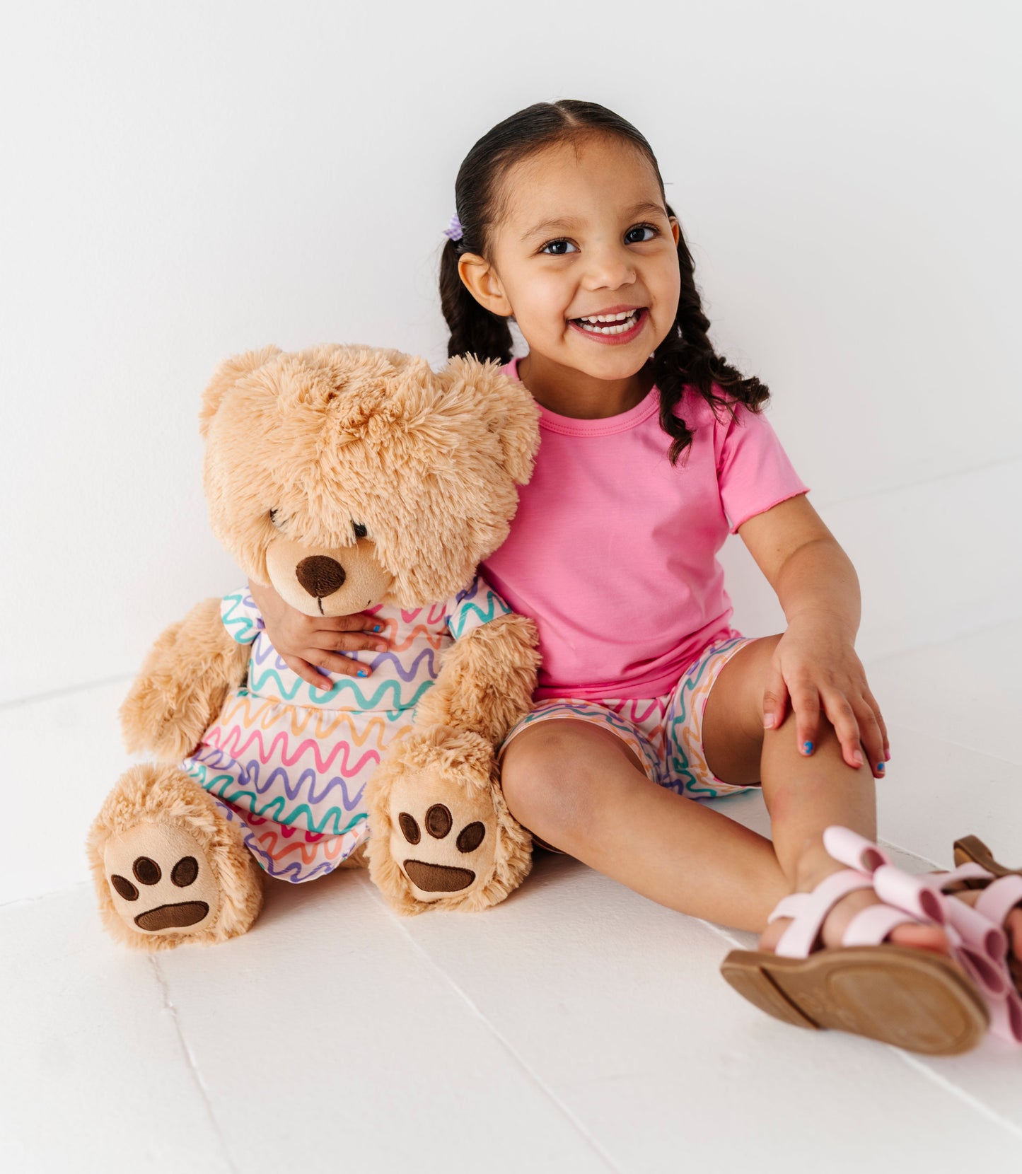 Child sitting on a white surface with a teddy bear wearing a colorful outfit.