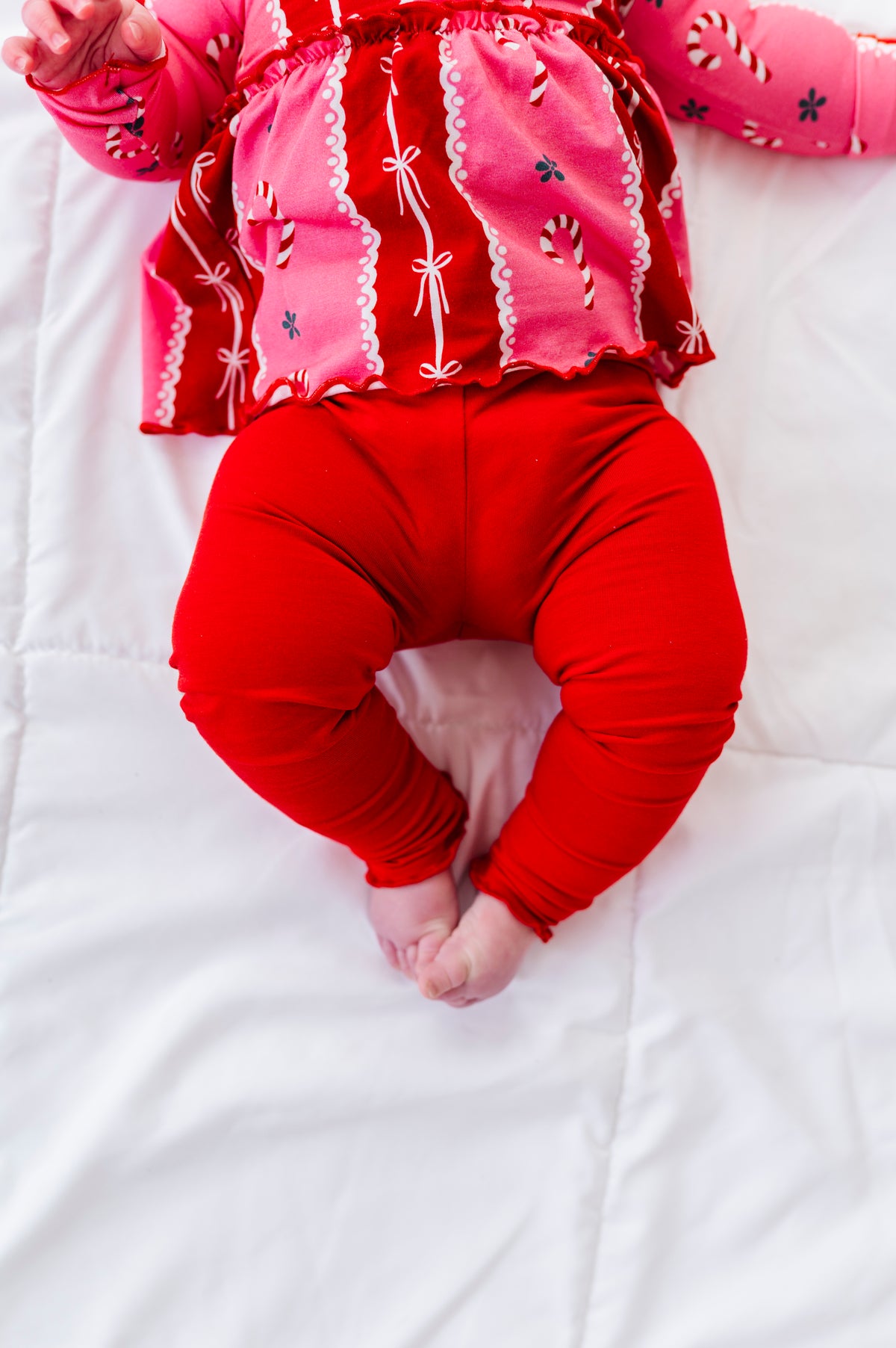 A baby lying on a white quilted blanket, wearing a bright pink long-sleeve bamboo top with white lace trim and candy cane motifs, paired with red leggings.