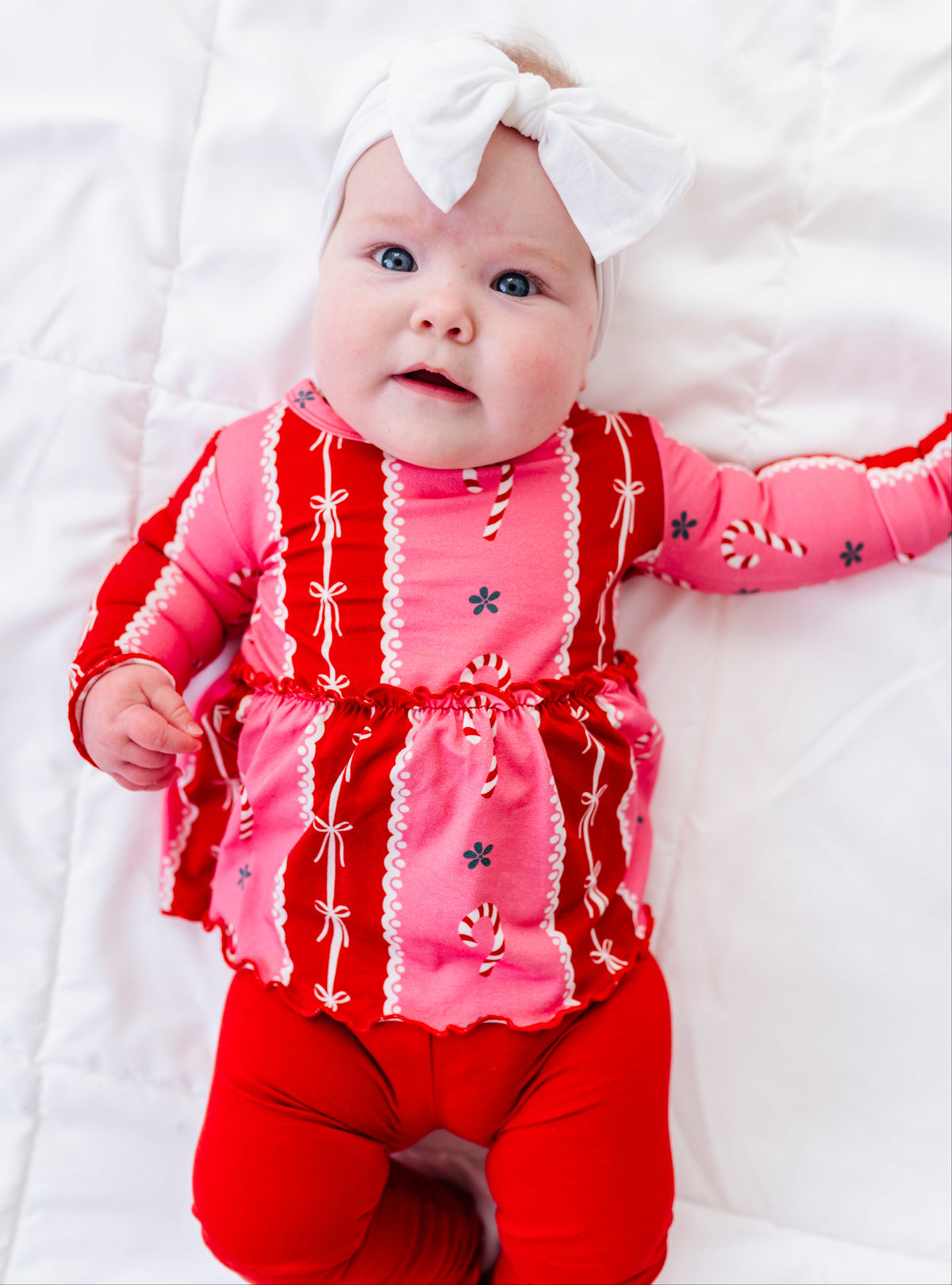 A baby lying on a white quilted blanket, wearing a festive holiday-themed two-piece bamboo outfit with a pink and red long-sleeve top featuring candy cane and snowflake patterns, matching red pants, and a white bow headband.