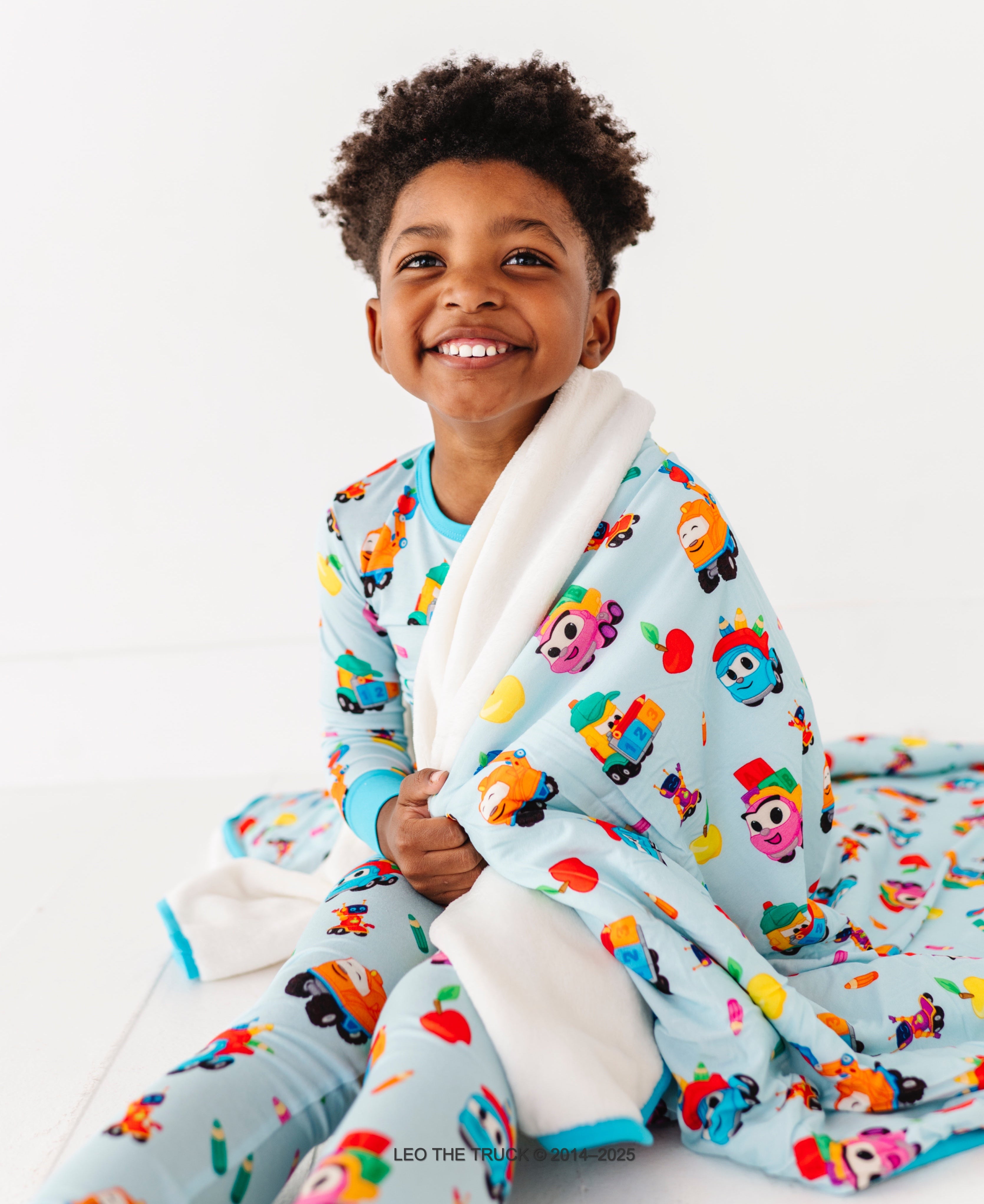 Young boy wearing colorful Leo the Truck bamboo pajamas and a matching blanket on a white background