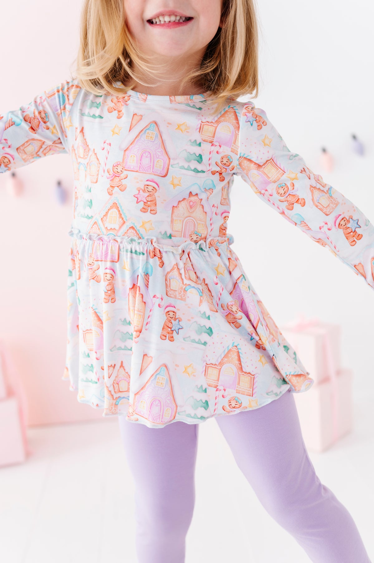 A little girl wearing a Christmas gingerbread-themed bamboo peplum top with purple leggings. She's standing in front of pink presents and a pastel Christmas lights banner. 