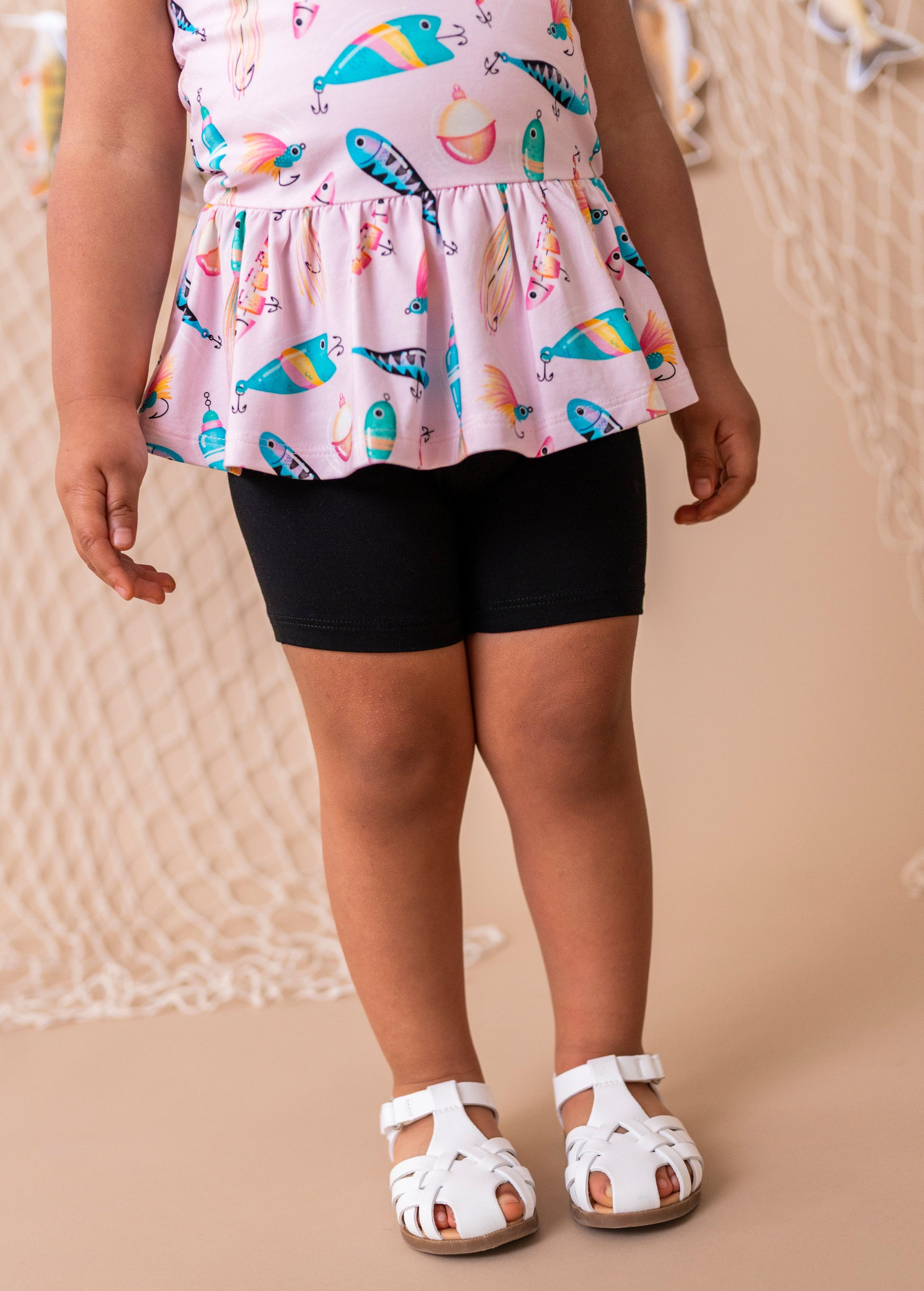 Child wearing a colorful top with fish-themed pattern and black shorts on a beige background