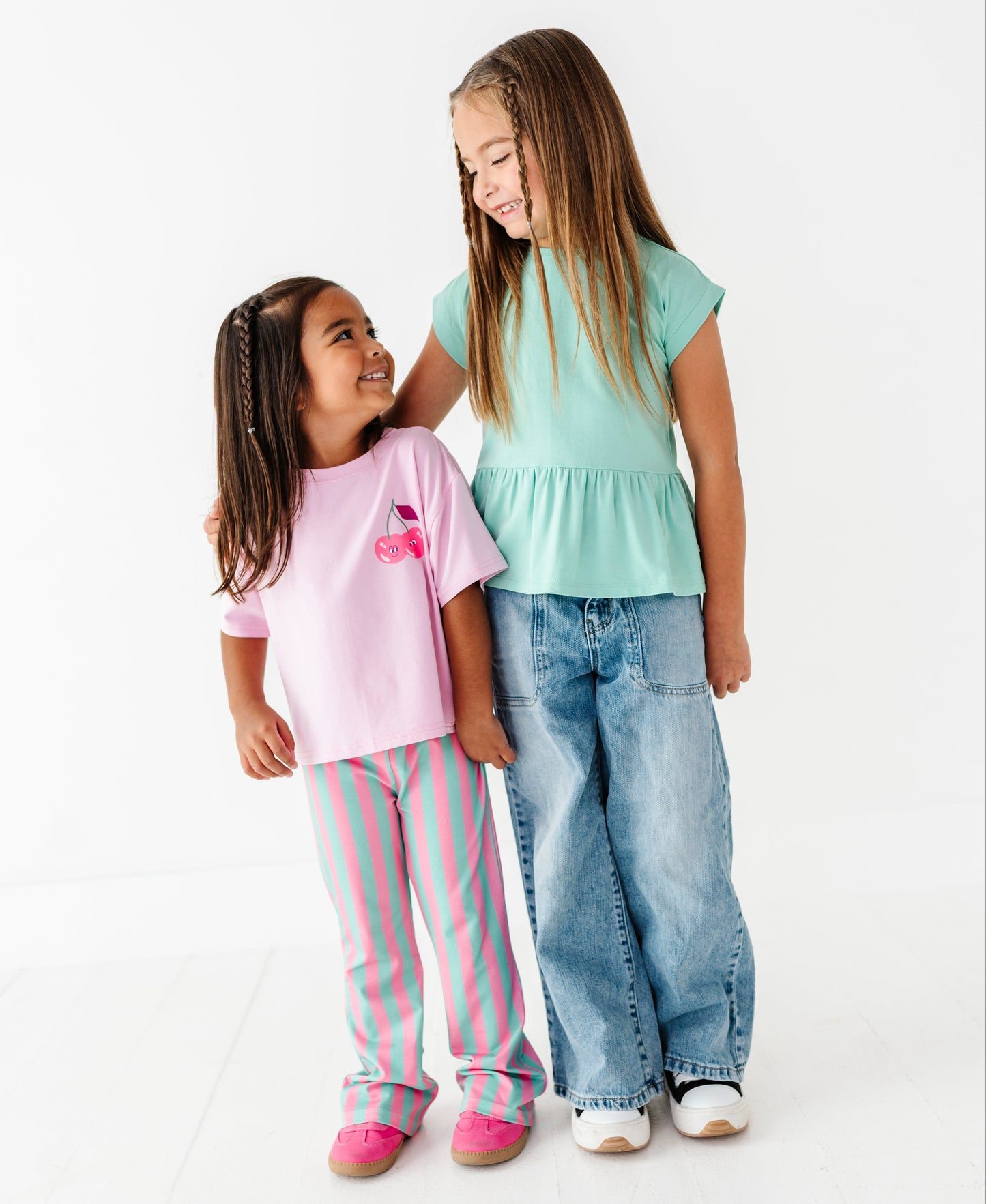 Two young girls standing together against a clean white studio background. The girl on the left wears a light pink short-sleeve top with a small cherry graphic and pink-and-white striped pants.