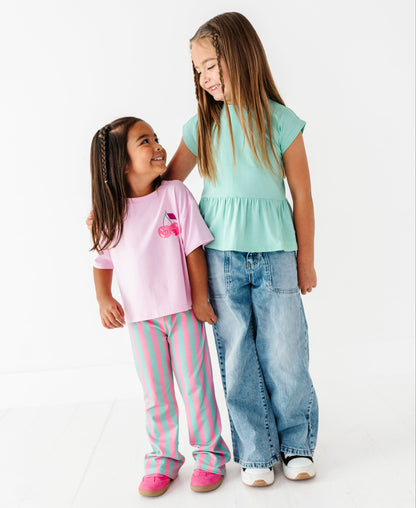 Two young girls standing together against a clean white studio background. The girl on the left wears a light pink short-sleeve top with a small cherry graphic and pink-and-white striped pants.