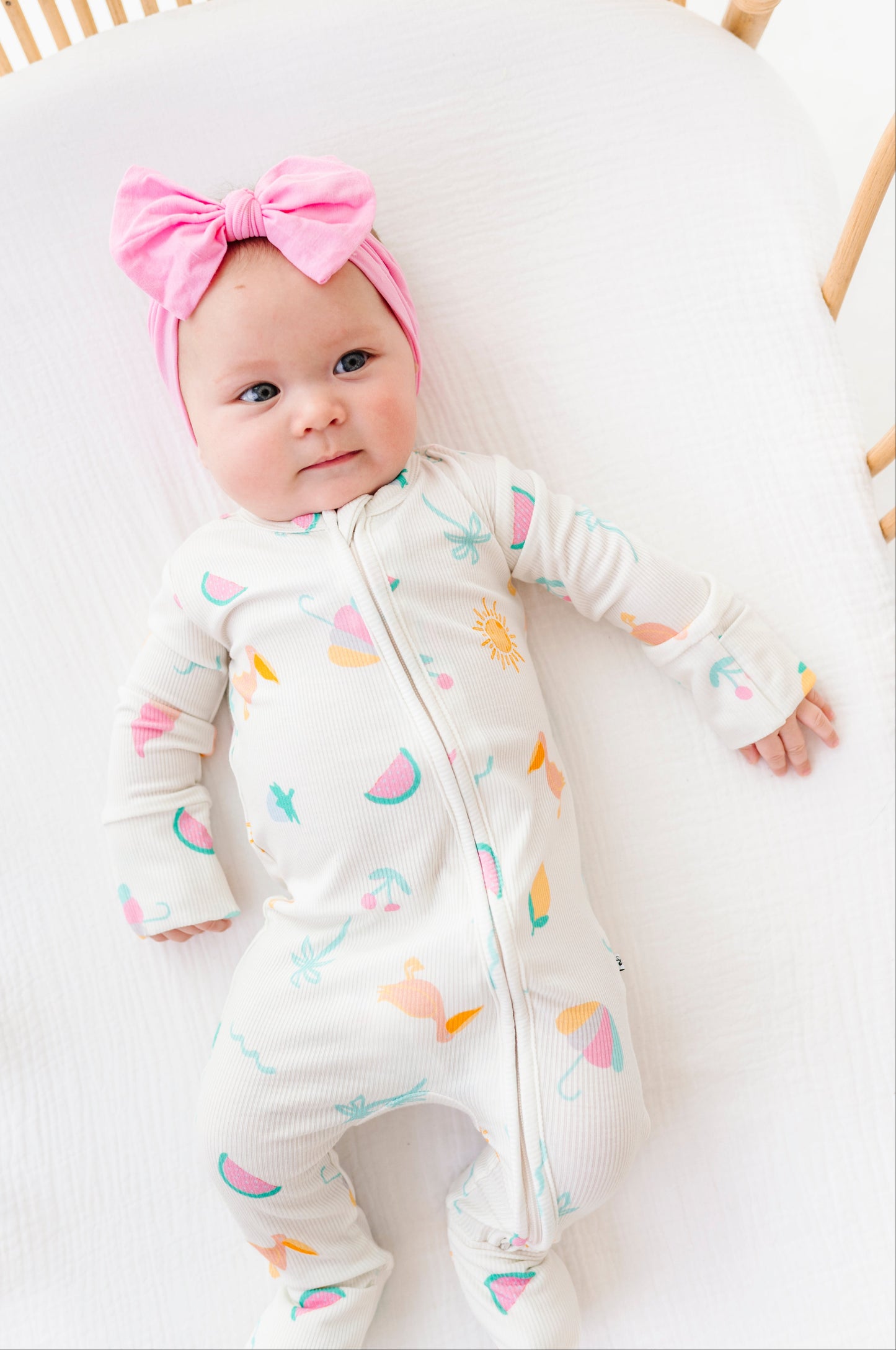 Baby in a colorful romper with a pink bow headband, lying on a white surface.