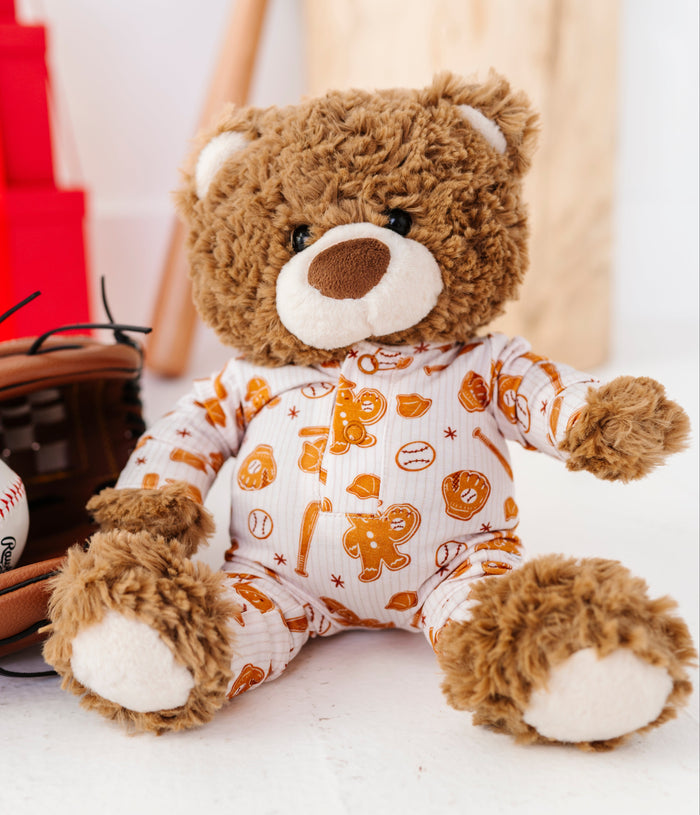 A brown plush teddy bear wearing a white long-sleeve pajama top with orange baseball-themed graphics, posed in a styled indoor setting with baseball props and a wooden cylinder background.