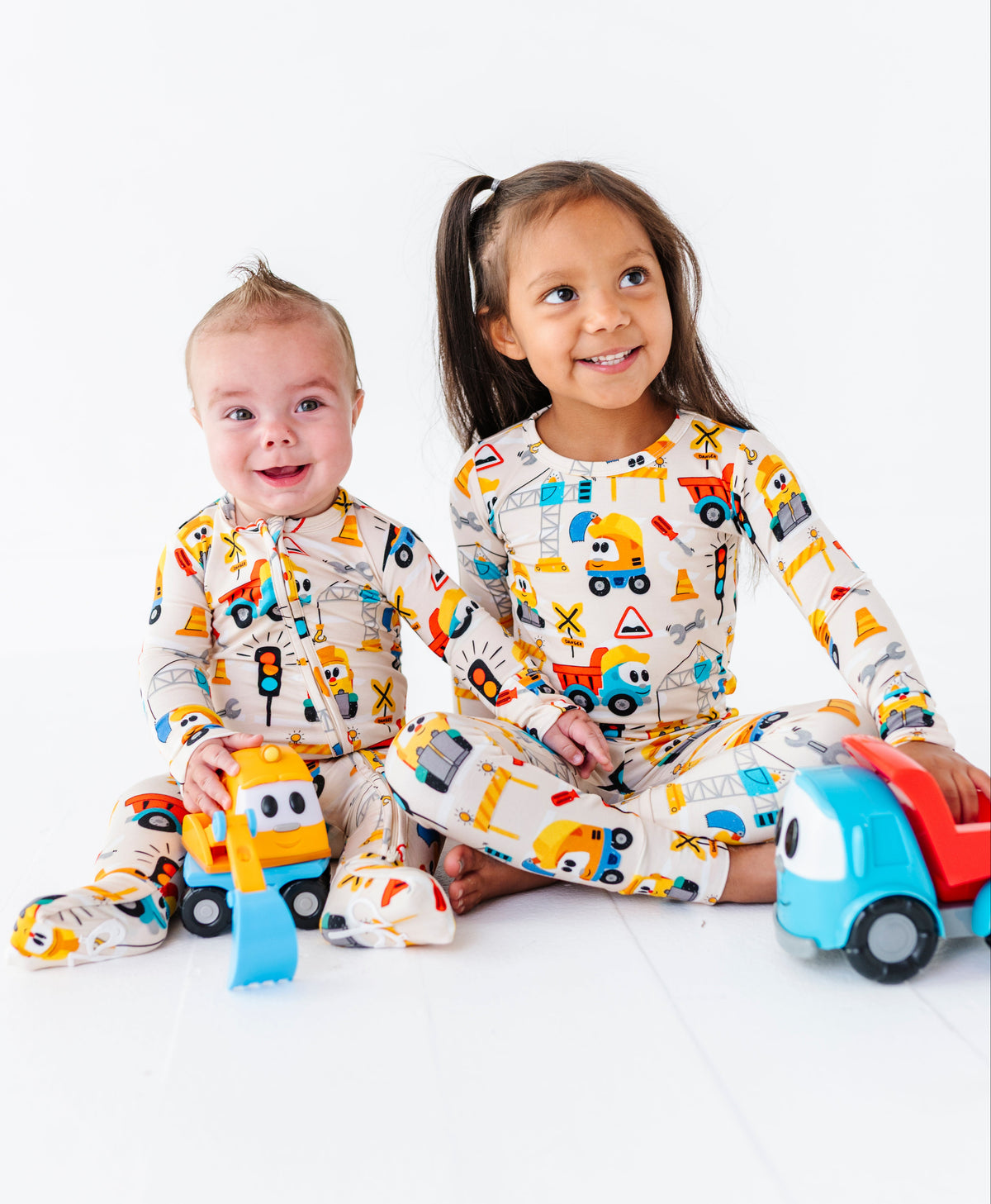 Two children sitting on a white floor wearing matching construction-themed pajamas, with toy vehicles in front of them.