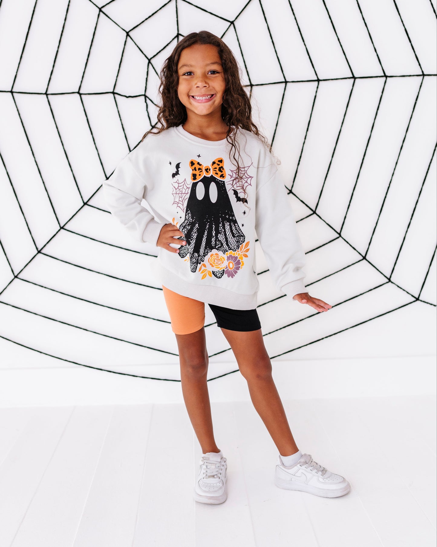 A young girl stands in front of a large black spiderweb graphic on a white background, wearing a white long-sleeve sweatshirt with a black ghost and orange spider graphic, black bike shorts, and white sneakers.