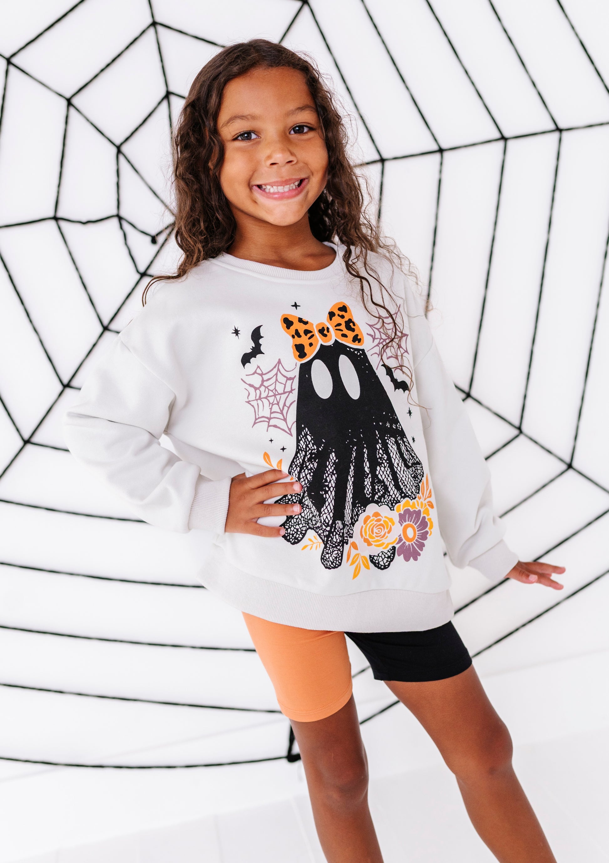A young girl with long brown hair smiling, wearing a white long-sleeve sweatshirt with a black ghost, orange bow, and spiderweb graphic. She is posing in front of a white background with black spiderweb line art.