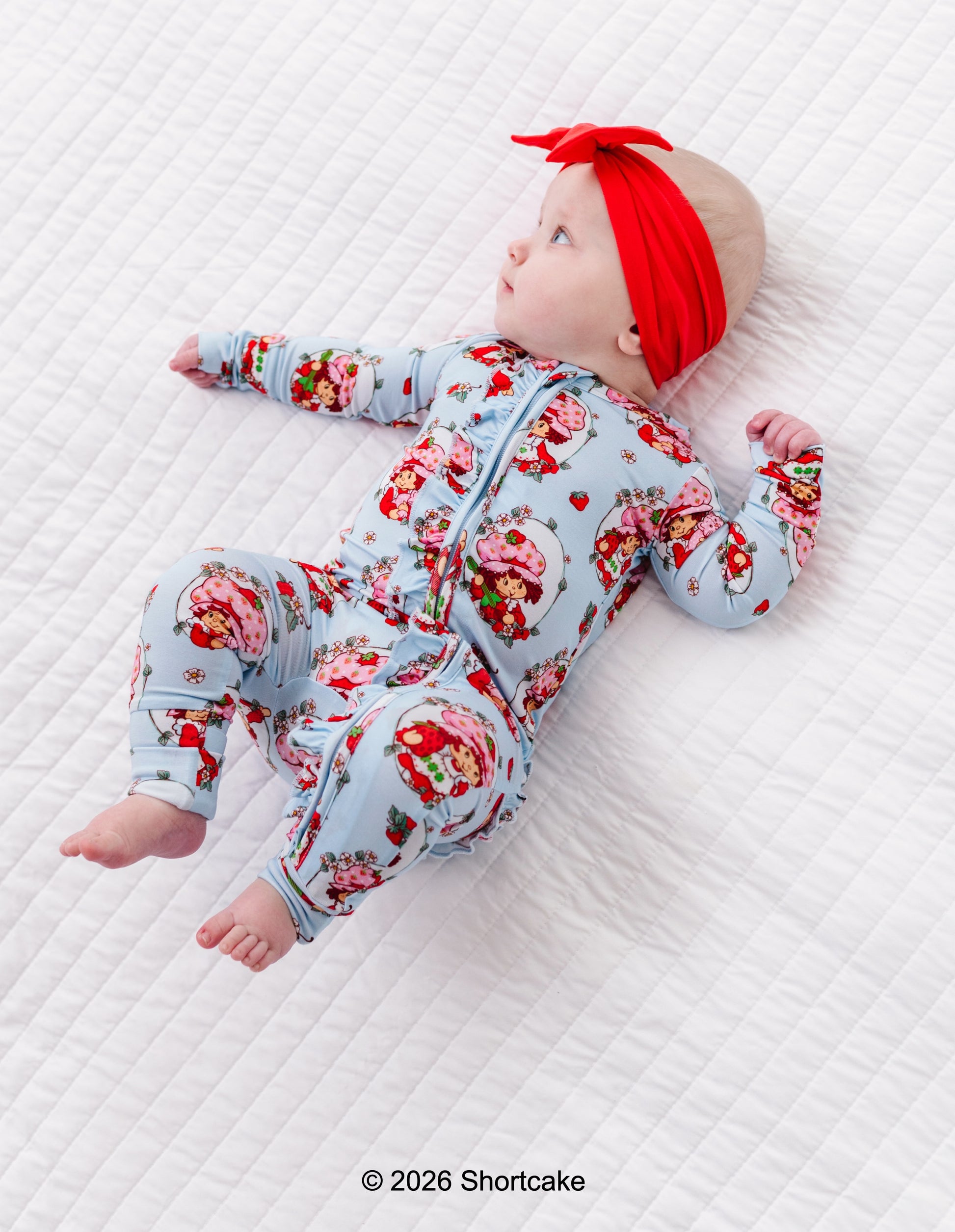 Baby in Strawberry Shortcake™ convertible romper with red headband lying on a white textured surface