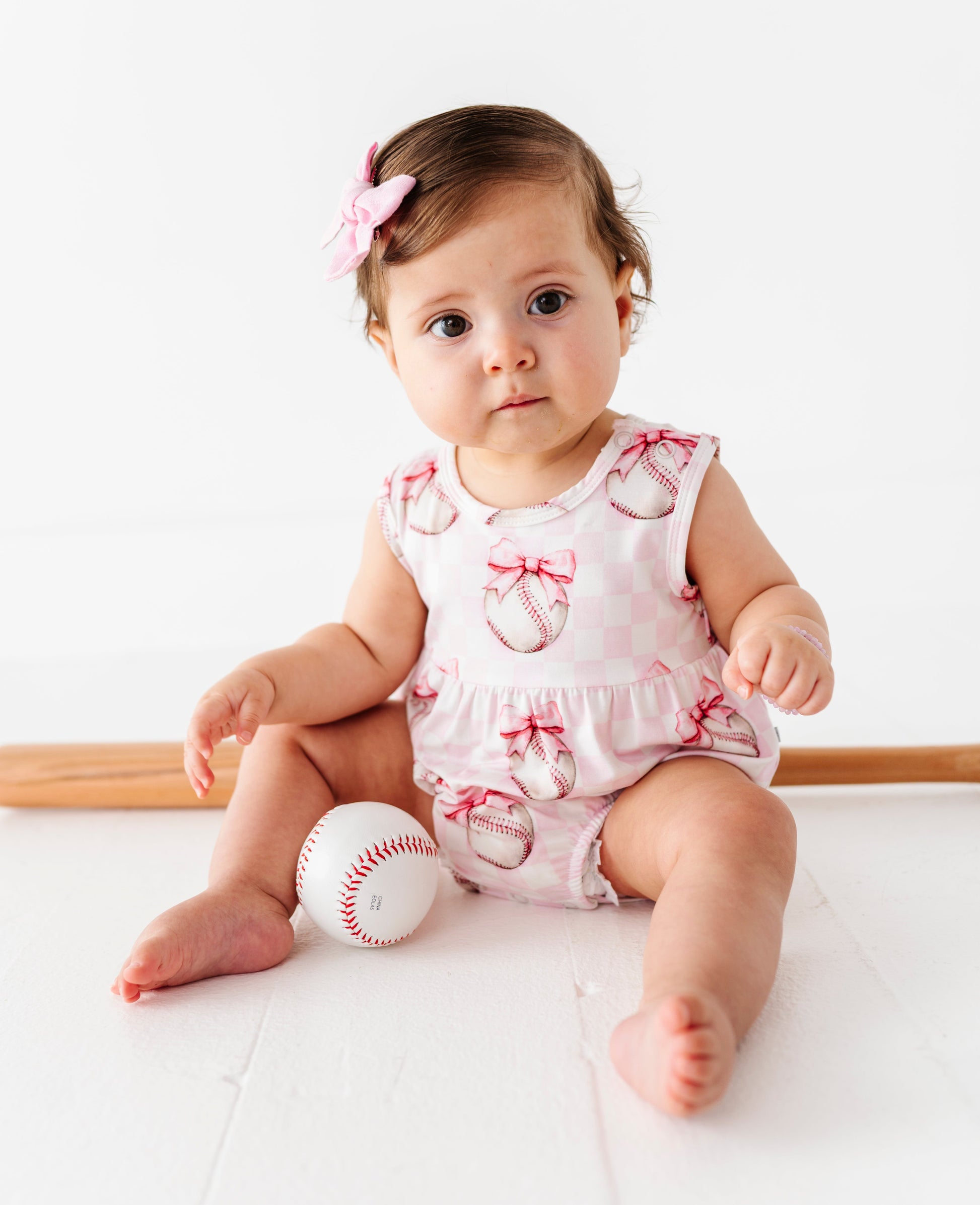 Baby in a pink bamboo outfit with baseballs and bows sitting on a white surface with a white background