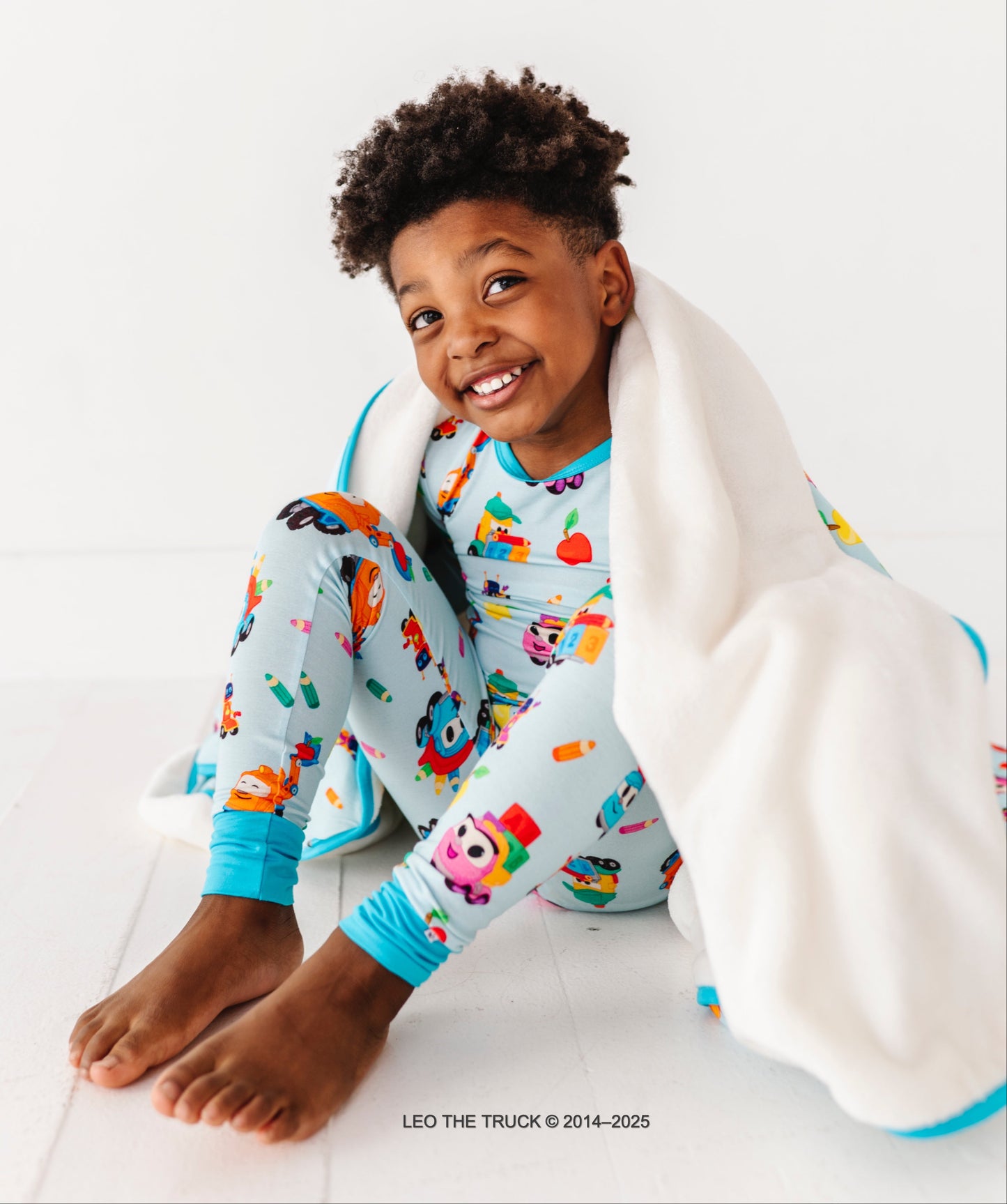 Toddler boy wearing blue Leo the Truck two-piece pajamas and wrapped in a plush bamboo Leo the Truck blanket 