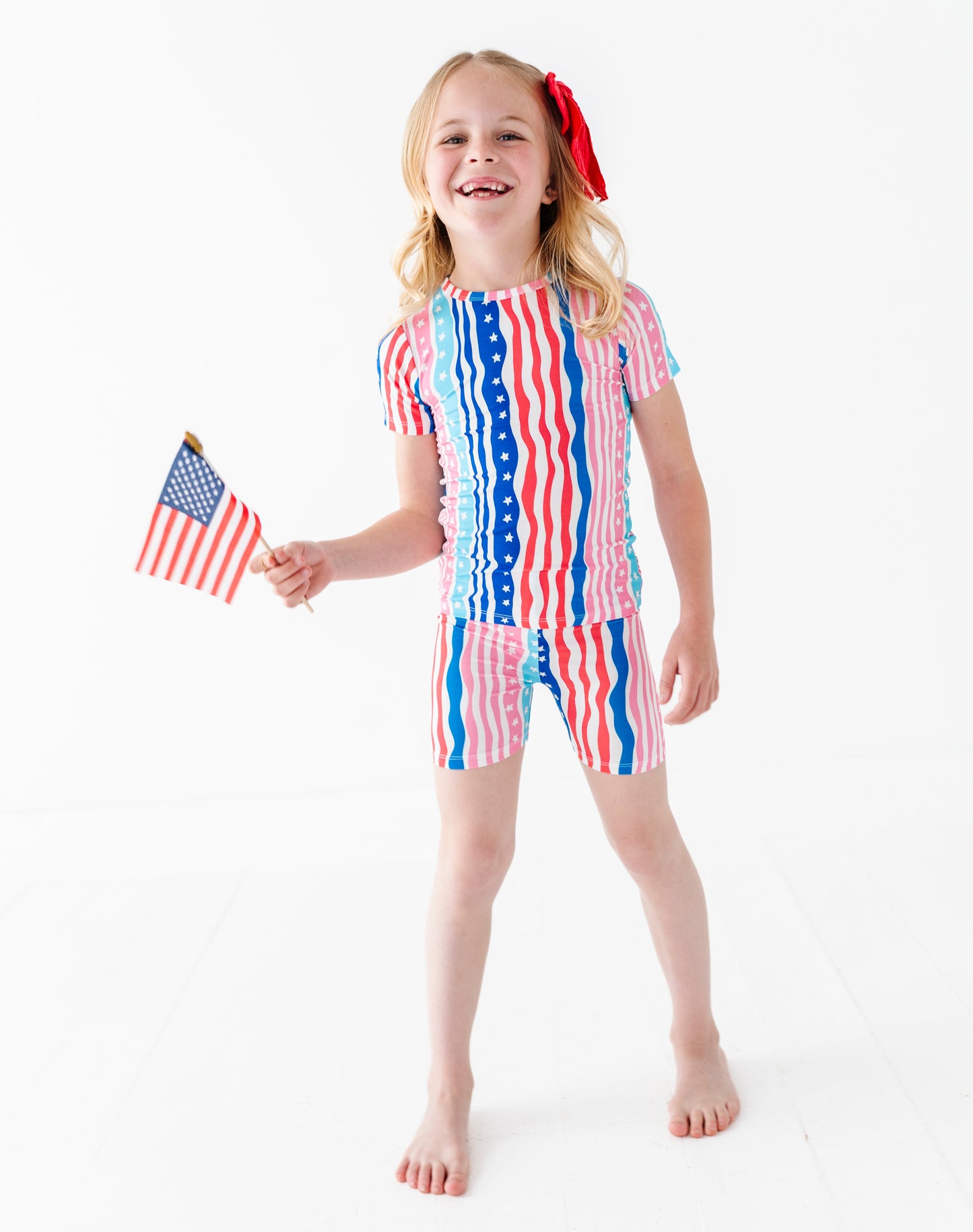 Young girl wearing a red, white, and blue striped bamboo pajama short set holding an American flag on a white background
