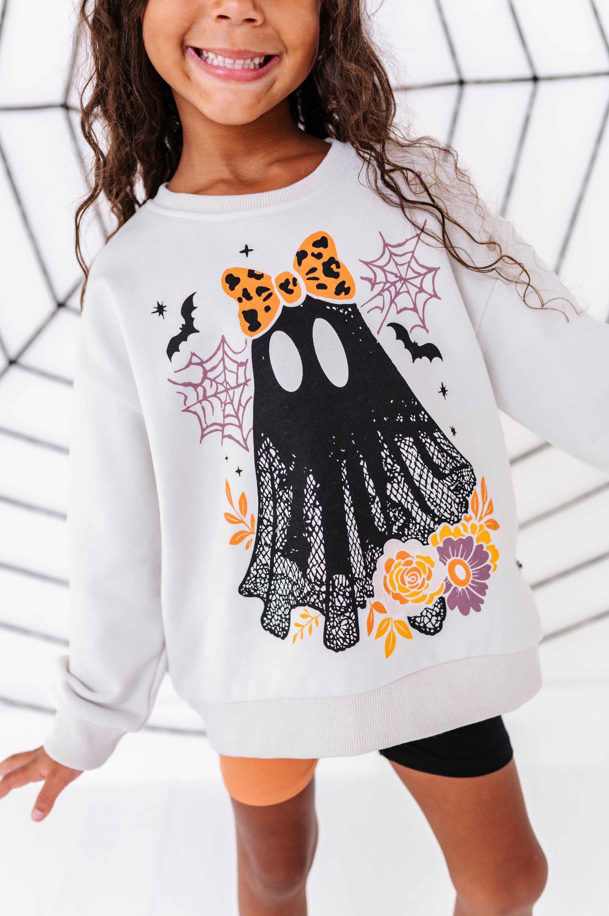 A child wearing a white long-sleeve sweatshirt with a Halloween ghost graphic featuring a black lace-patterned ghost, orange leopard-print bow, orange and yellow flowers, and bats, posed against a white web-patterned backdrop.